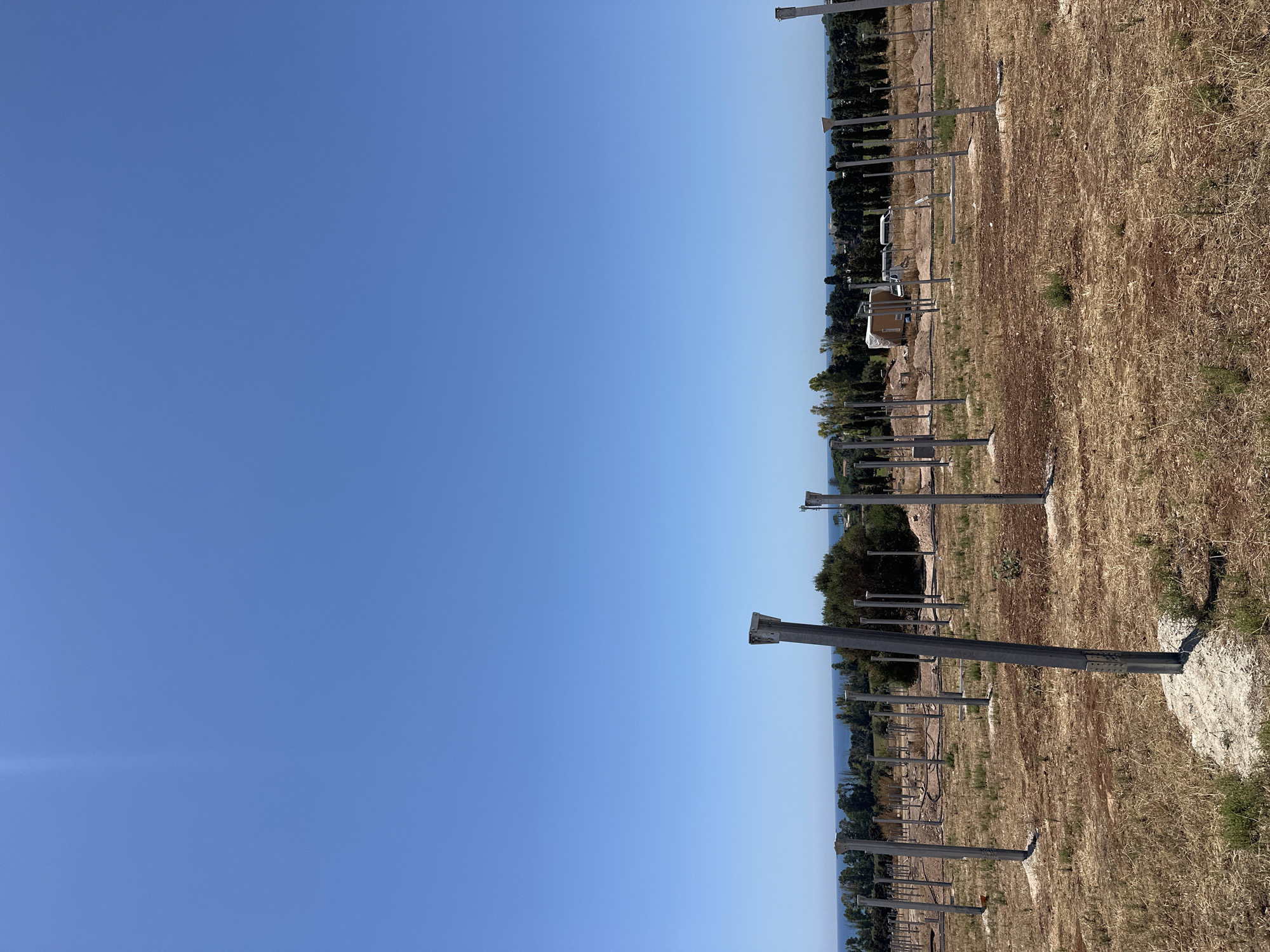 The image depicts an outdoor scene under a clear blue sky. In the foreground, a dry, grassy field is dotted with numerous tall, slender metal posts, many of which appear to be part of a construction or installation project. Some posts are topped with rectangular fixtures. The ground is dry and reddish-brown, with sparse vegetation. In the middle ground, there are more rows of these posts, along with some low-lying structures, possibly portable buildings or equipment, and what appears to be a white van. Beyond this area, a line of trees forms a backdrop, with the blue sea visible in the distance. The overall impression is of a site under development, likely in a rural or coastal area.
Based on the context provided in the prompt, this scene is located in the city of Scicli, Italy.