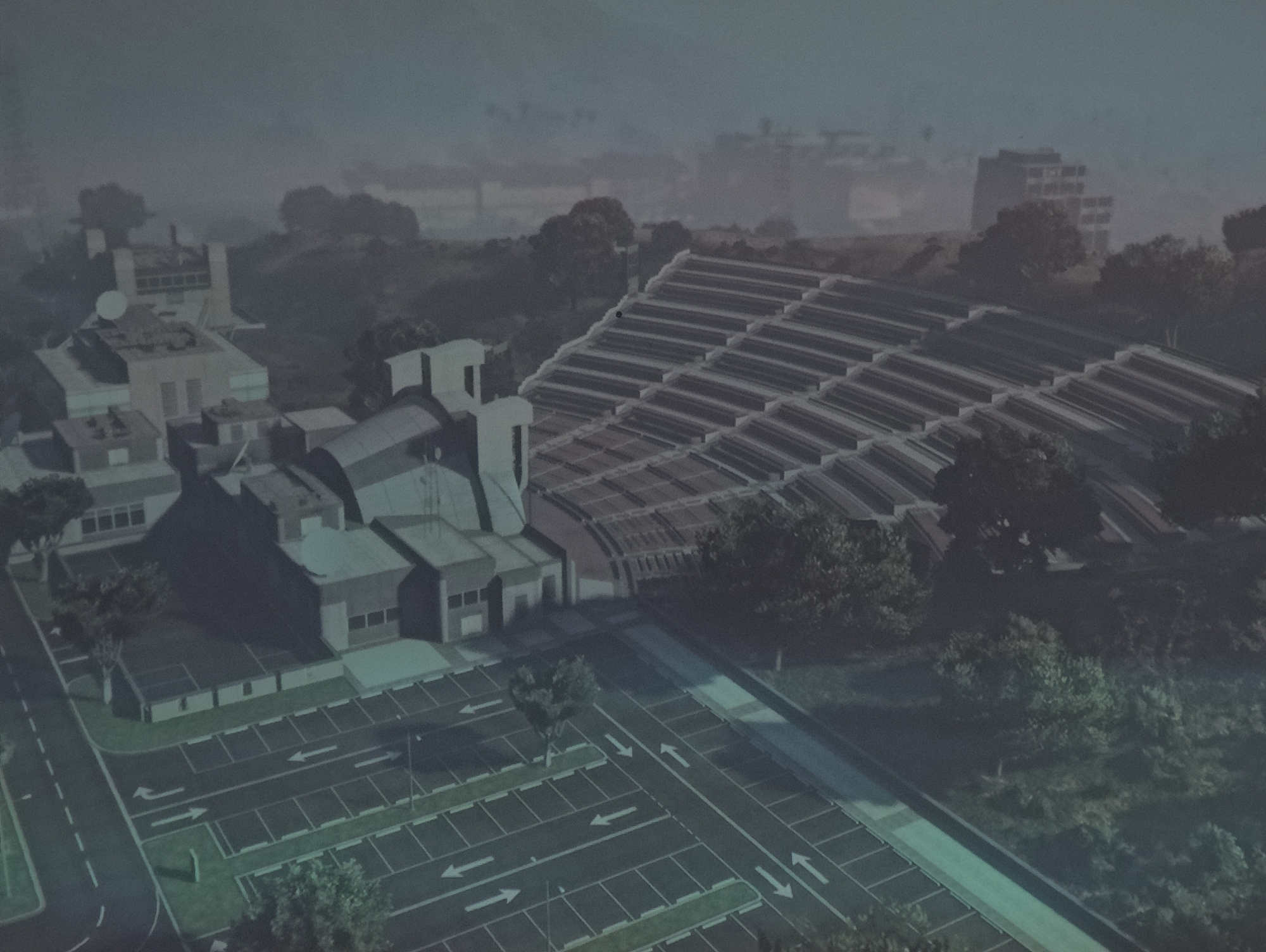The image presents an aerial view of a complex in East Suffolk, United Kingdom. It features a cluster of modern, geometric buildings with flat roofs and a light-colored facade. One prominent structure is topped with a satellite dish. Adjacent to the main buildings is a large, sloping structure comprised of multiple parallel rows, resembling a stadium or greenhouse. A large parking lot with clearly marked spaces and directional arrows is visible in the foreground. A road runs alongside the parking lot and connects it to the complex. The scene is surrounded by trees and greenery, with some buildings visible in the distant background. The overall color palette is muted, with a predominance of gray and green hues. The scene appears to be shrouded in a light mist or fog, giving it a slightly hazy appearance.