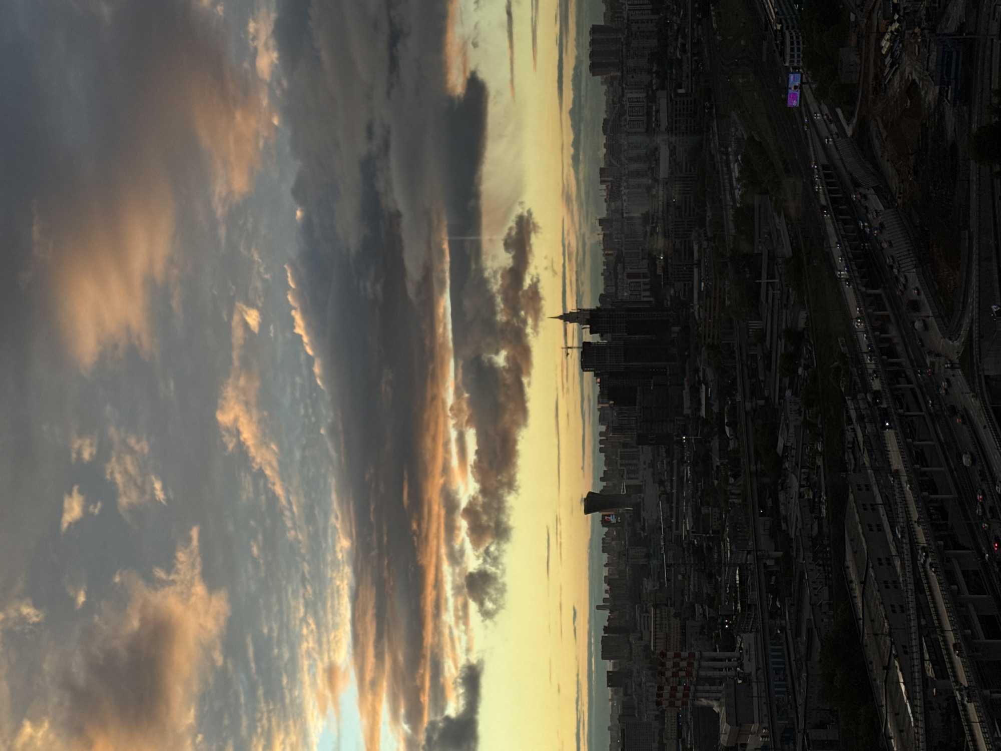 The image depicts an urban environment, likely the city of Khimki in Russia, viewed from a high vantage point during twilight or sunset. The sky occupies a significant portion of the frame, showcasing dramatic cloud formations illuminated by the setting sun, creating a gradient of colors from blue to orange and grey. Below the horizon, a dense cityscape is visible, composed of numerous buildings of varying heights. Some structures appear to be high-rise apartments or office buildings. Several major roadways with moving vehicles can be seen, suggesting a busy transportation network. The image has been rotated 90 degrees clockwise.
