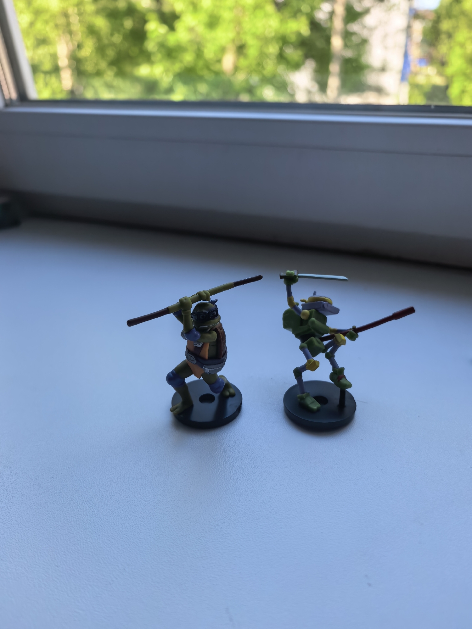 The image shows two miniature Teenage Mutant Ninja Turtles figures standing on a white surface. One figure is orange and wielding a bo staff. The other is green and holding a sword in one hand and a bo staff in the other. The figures are on black bases. The background includes a window with blurry green foliage visible outside. There is no evidence to suggest this photo was taken in Sterlitamak, Russia.