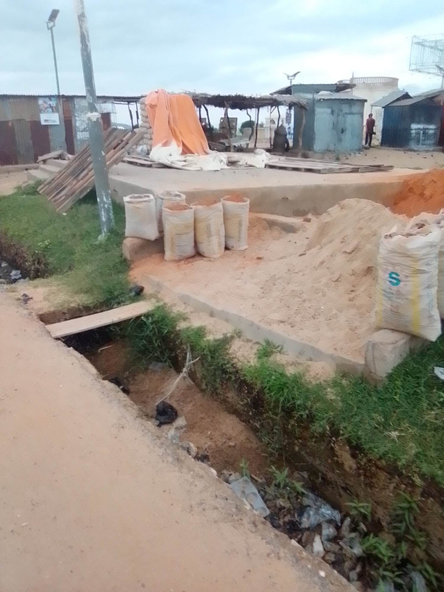 The image depicts a street scene in Kwadon, Nigeria, characterized by a dirt road and a roadside drainage ditch. Several large sacks, presumably filled with sand or cement, are arranged on a raised concrete platform. In the background, various structures are visible, including corrugated metal sheds and a covered market stall draped with an orange tarp. A wooden pallet lies near some of the sacks. The drainage ditch contains some debris, including plastic bags and what appears to be dark material, possibly refuse. The sky is overcast. A single person is visible in the distance, standing near one of the structures. A street lamp is also present on a pole in the foreground. The overall impression is one of a developing or semi-built area.