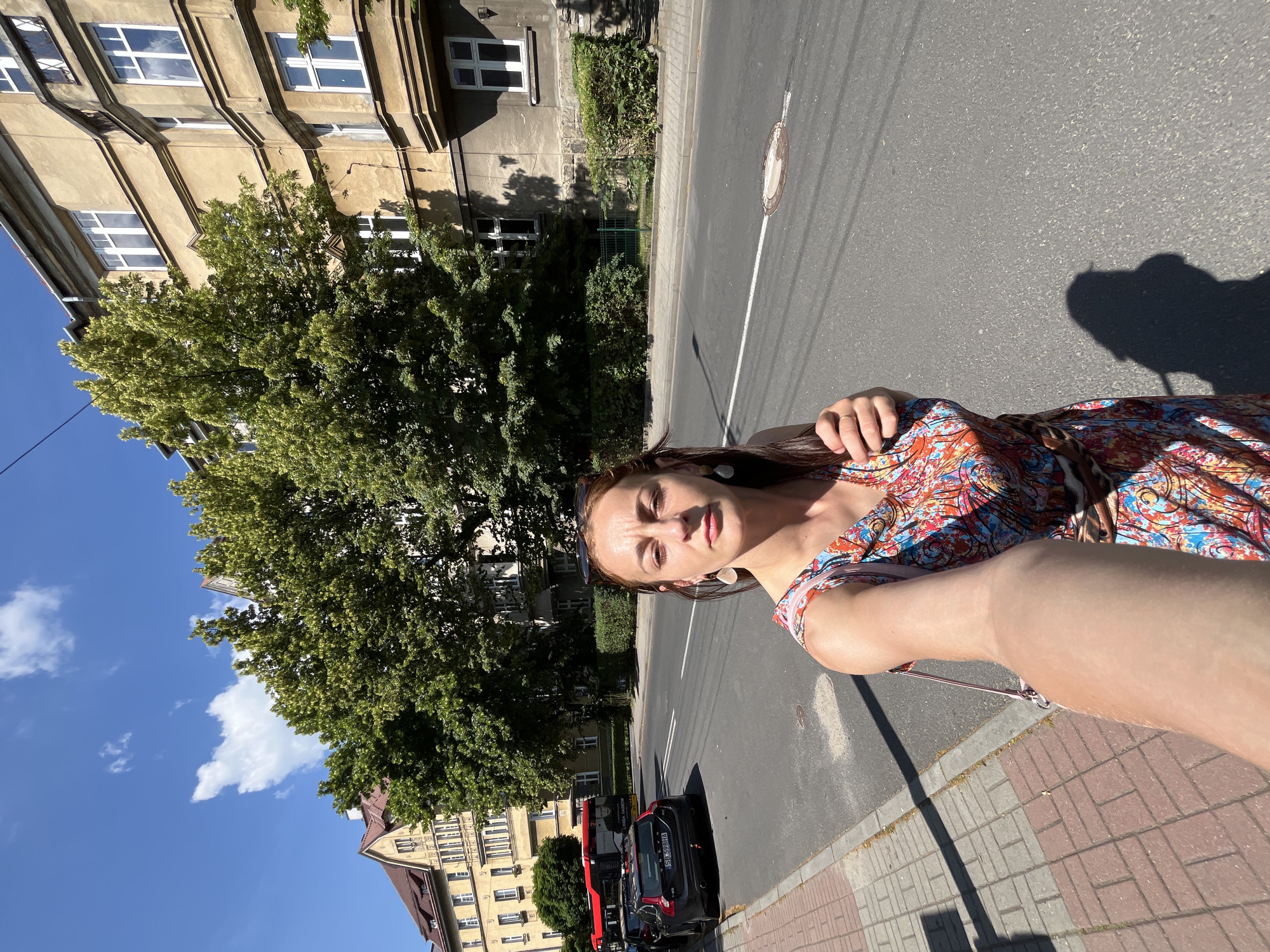 This is a selfie taken by a woman standing on a sidewalk next to a street. She is wearing a brightly colored, patterned dress. The background shows a building with several windows, a large green tree, and a clear blue sky with some clouds. There is a street with a white line, a manhole cover, and parked cars. A shadow of the woman and possibly something she is holding is cast on the street. The image appears to be taken during the daytime with bright sunlight, as indicated by the strong shadows. The woman's expression is neutral. There is a license plate visible on a black car that reads "SK 598IA".