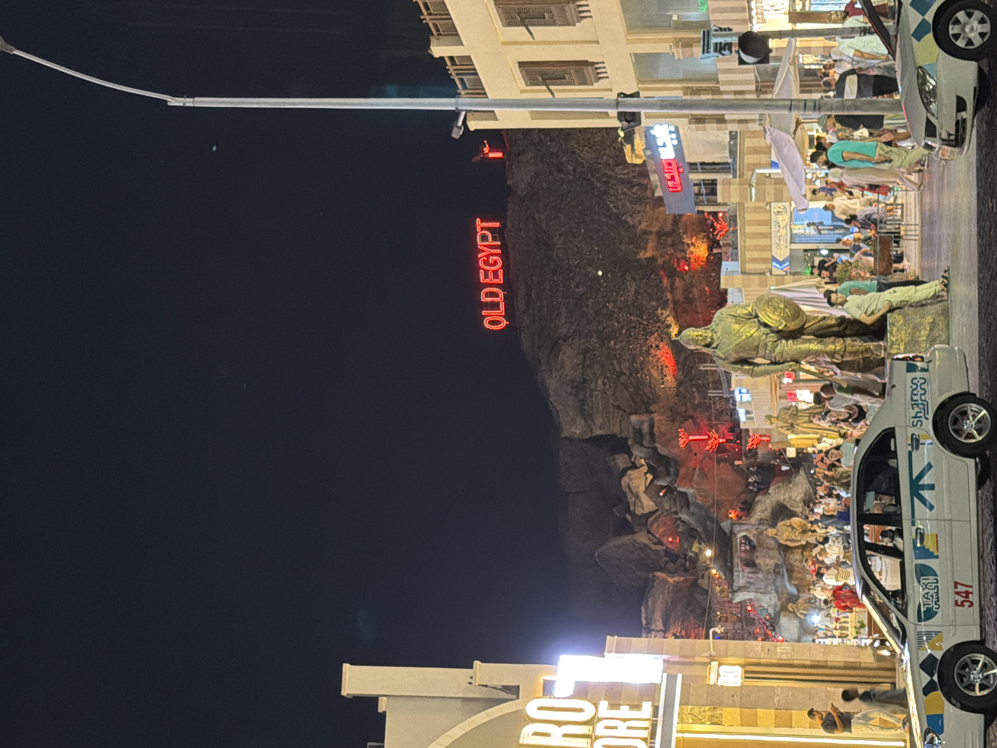 The image captures a bustling nighttime scene in Sharm El Sheikh, Egypt. A prominent neon sign reading "OLD EGYPT" is illuminated in bright red atop a rocky hill, serving as a backdrop. In the foreground, a white taxi with the number "547" visible on its side is parked on the street. A statue of a man in historical attire, holding a shield, stands to the left of the taxi. The street is filled with a crowd of people, some appearing to be tourists, mingling around shops and stalls. The architecture suggests a commercial or entertainment district, with buildings adorned with traditional elements. Red lights are visible throughout the scene, contributing to the vibrant atmosphere. The overall impression is one of a lively tourist destination at night.