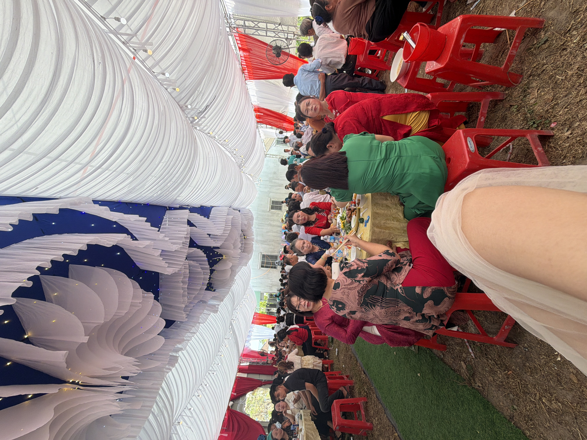The image shows an outdoor event in Tay Son Commune, Vietnam. Many people are seated at tables under a large tent structure decorated with white fabric draping and blue sections. The tent also features some lighting elements. People are dressed in various clothing, suggesting a social gathering such as a wedding or a party. Red plastic stools are used as seating. The ground is a mixture of grass and bare earth.