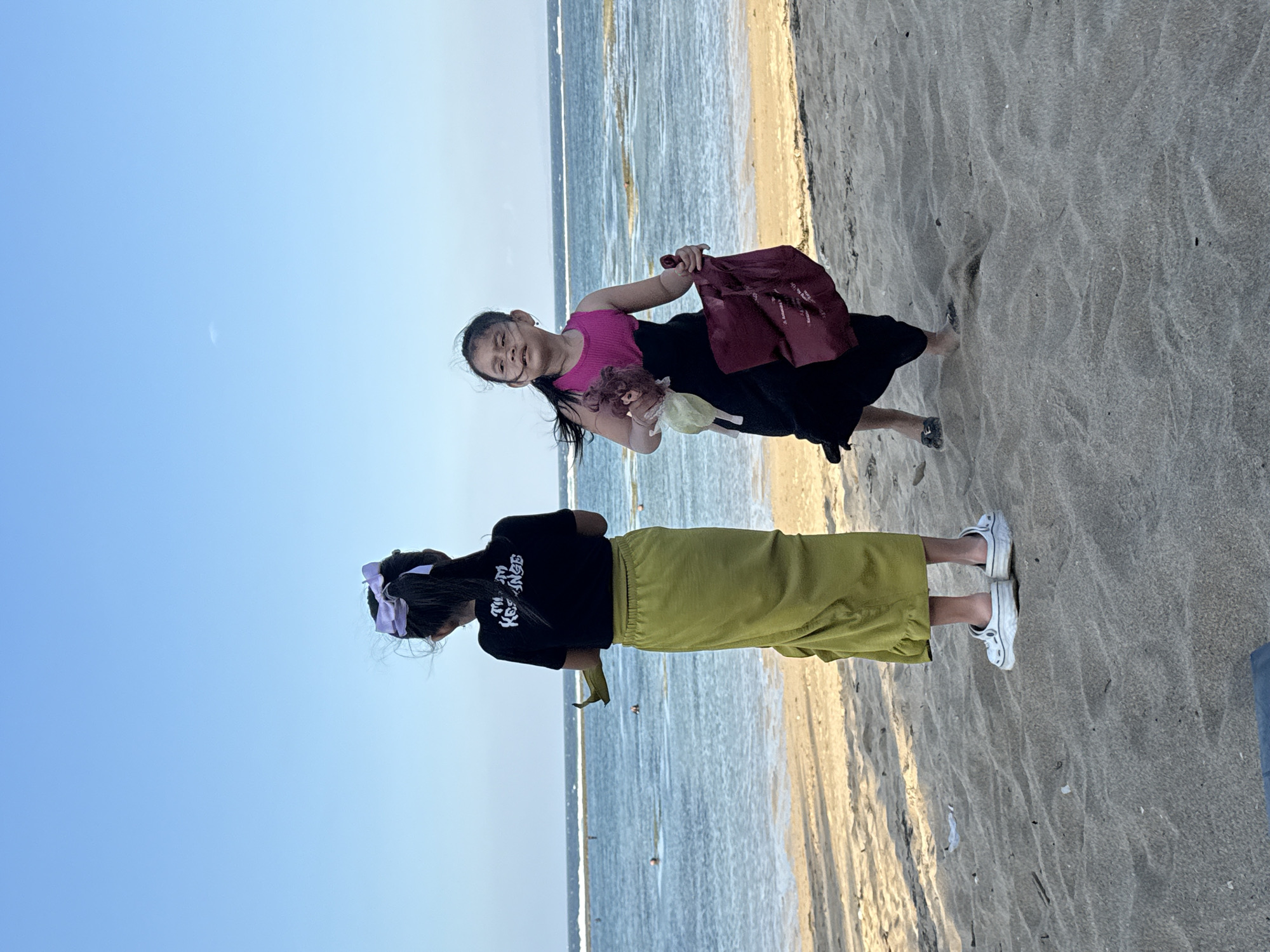 Two young girls are standing on a sandy beach in Denpasar, Indonesia, facing the ocean. The girl on the left, facing away from the camera, wears a black t-shirt with white lettering and a long, lime green skirt. She has a purple bow in her hair. The girl on the right, facing the camera, wears a pink and black dress. She is holding a doll in her left arm and a dark red bag in her right hand. The ocean stretches out behind them, with small waves breaking near the shore. The sky is clear and blue. The sand is light brown and shows footprints.