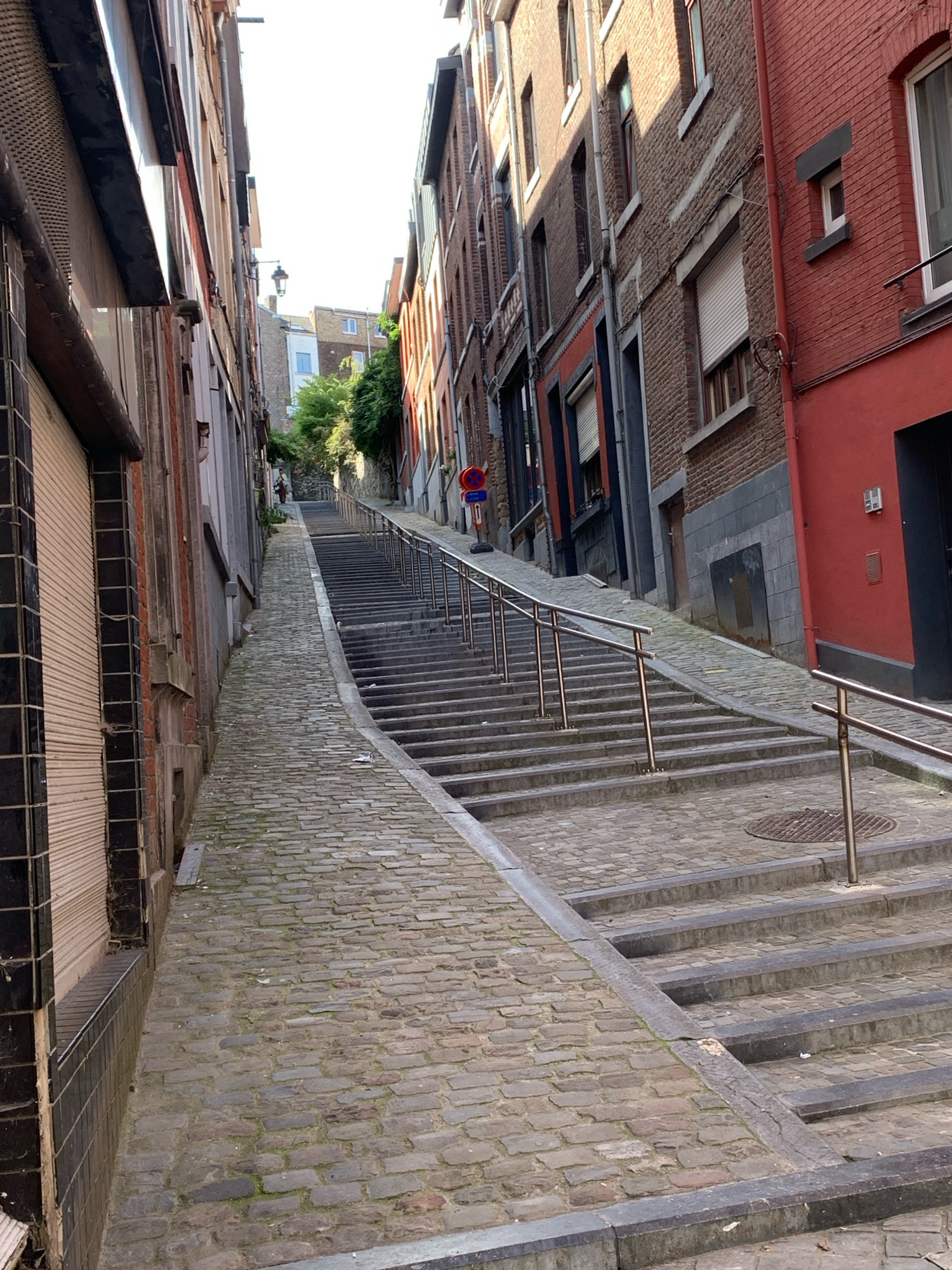 The image depicts a narrow cobblestone alleyway in Liège, Belgium, ascending a steep incline. The alley is flanked by tall, old brick buildings with a mix of architectural styles, some featuring exposed brick and others painted in vibrant colors like red. Metal railings line the right side of the alley, accompanying a series of stone steps that lead upwards. Further up the alley, a lone figure is visible walking away from the camera. A street lamp is also present, mounted on a pole. The sky is bright, suggesting daytime.