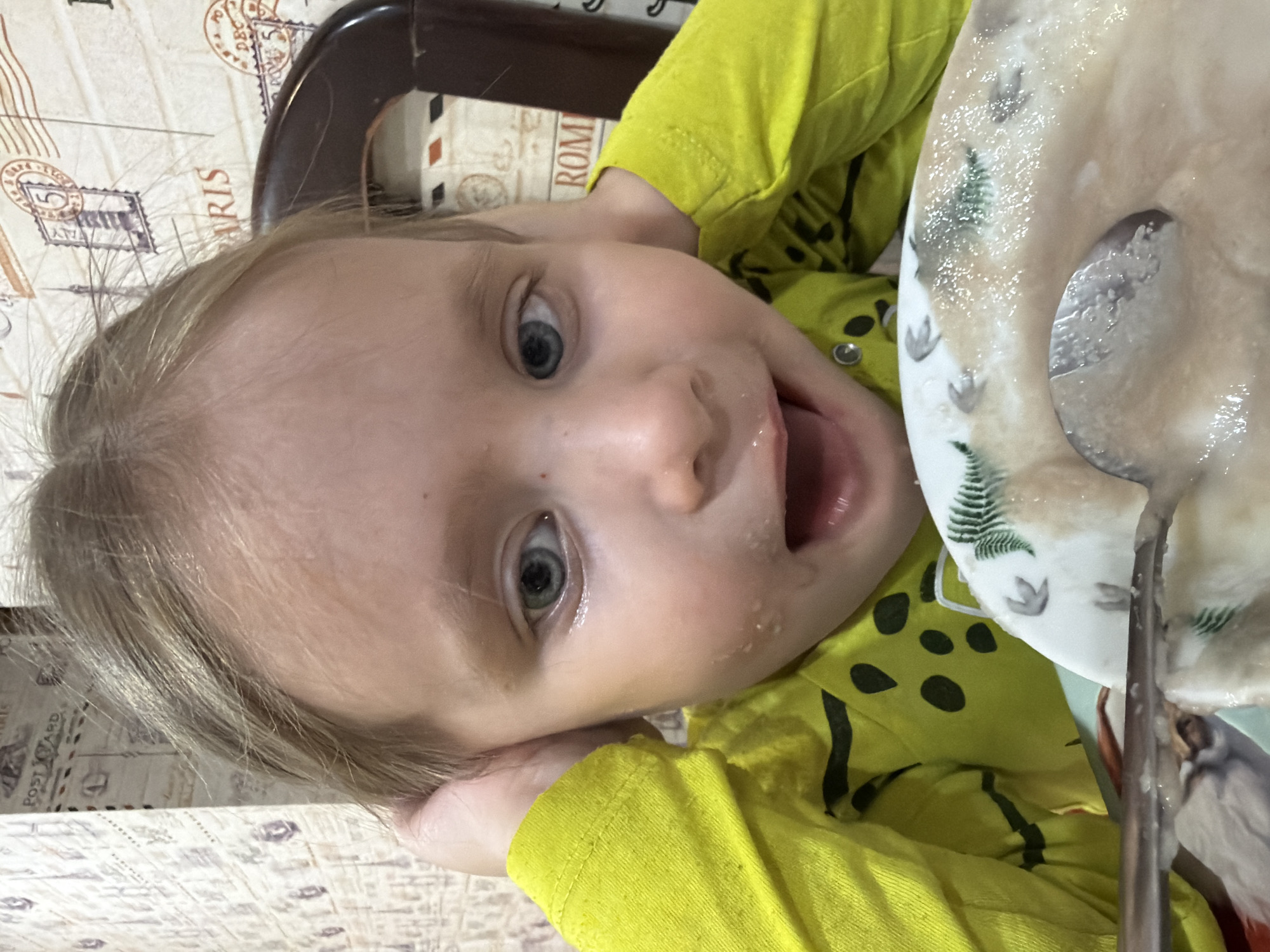 A child with blond hair and blue eyes is lying on their side at a table. They are wearing a bright yellow shirt with black spots. The child's mouth is open and there is food on their face, suggesting they are eating. A plate with food and a spoon are visible nearby. The table has a patterned surface resembling a collage of postcards or letters. The photograph was taken in Chaikovsky, Russia.