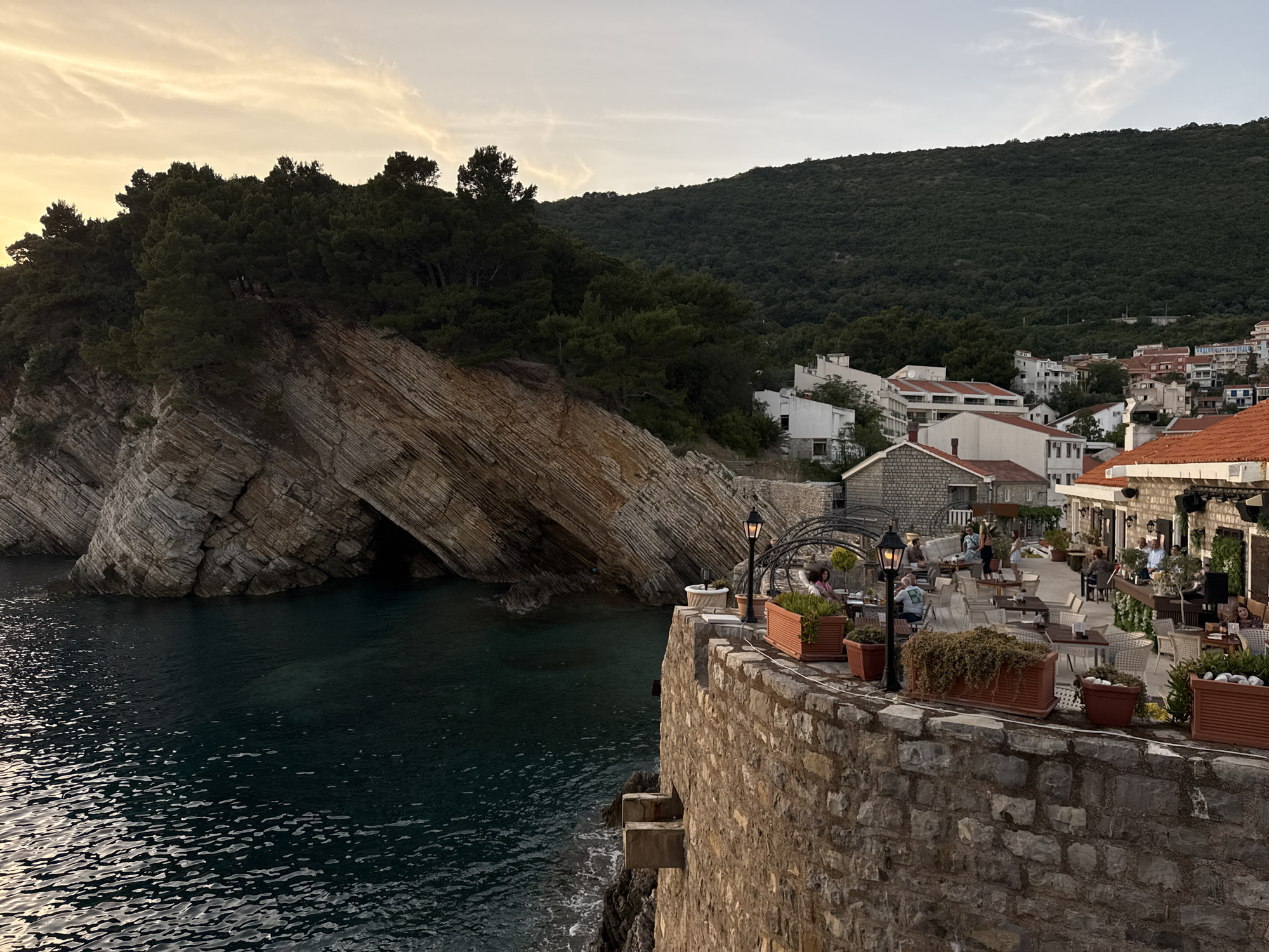 The image depicts an outdoor restaurant setting at the edge of the Adriatic Sea in Petrovac na Moru, Montenegro. A stone wall lined with planters filled with greenery borders the restaurant, which is furnished with tables and chairs occupied by patrons. Some are seated at tables, while others are standing. In the background, buildings with orange tiled roofs are visible, interspersed with green trees. Beyond the restaurant, a rock formation juts out into the water. The rock formation has a cave and trees at the top. The sea is calm and dark. The sky is overcast with soft light, suggesting either early morning or late afternoon.
