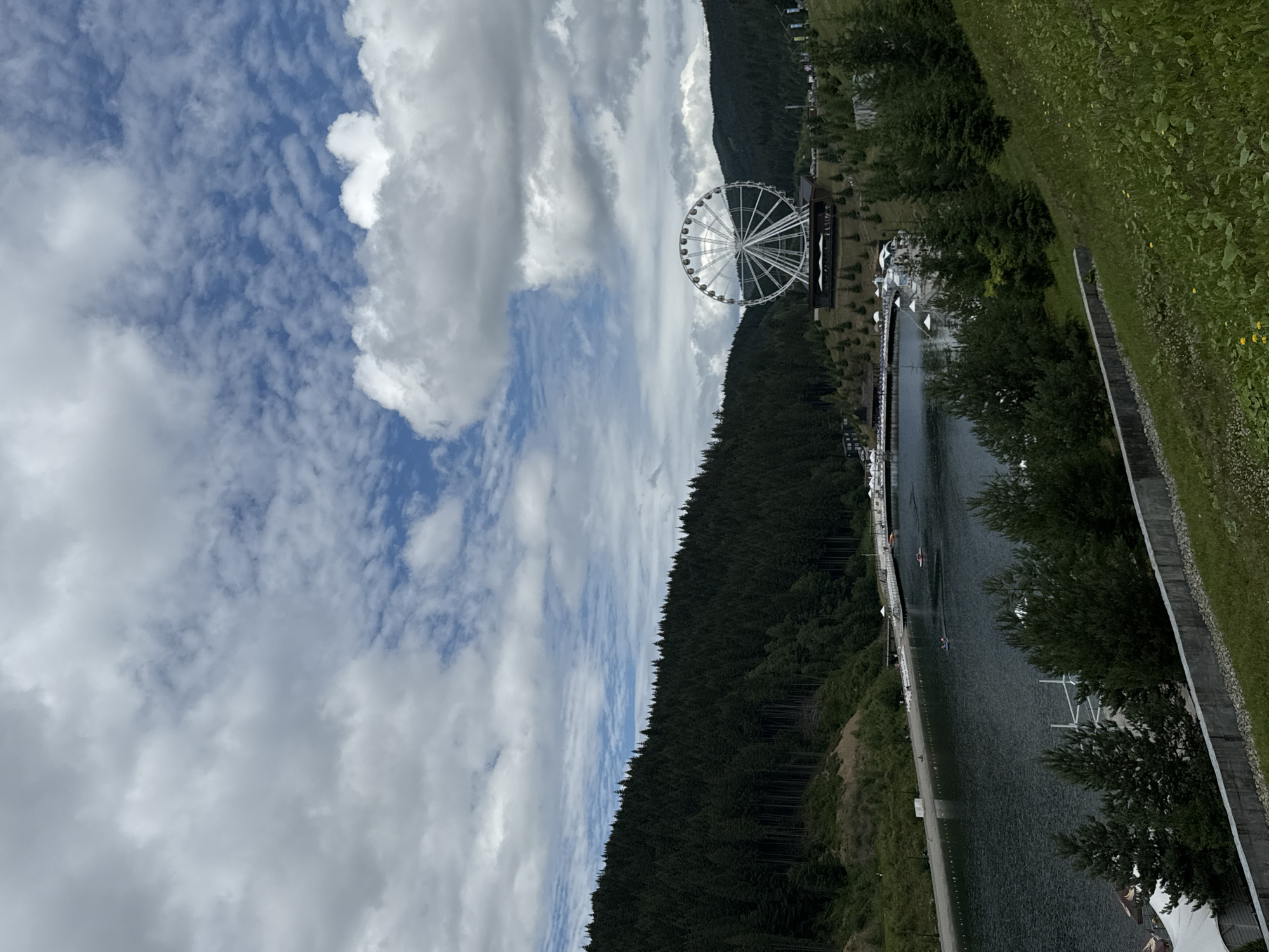 The image depicts a scenic view of a Ferris wheel situated in the city of Polianytsia, Ukraine, nestled amidst a picturesque landscape. The Ferris wheel, a prominent structure, is positioned on a hill overlooking a serene lake. The surrounding environment is characterized by lush green forests that ascend the slopes of the hills, creating a verdant backdrop. The sky above is adorned with a dynamic arrangement of clouds, with patches of blue sky peeking through.
In the foreground, a body of water, likely a lake, occupies a significant portion of the scene. The surface of the water is calm, with a few small boats visible, suggesting recreational activities. Along the edge of the lake, a sandy beach area is lined with lounge chairs and umbrellas, indicating a public recreational space. Further back, a pier extends into the water, with white structures that could be part of a boating facility.
The Ferris wheel itself is large and white, with numerous spokes and cabins, suggesting it is a significant attraction. It stands on a raised platform that features a building or structure beneath it, possibly a restaurant or viewing area. The Ferris wheel is surrounded by scattered trees, with more dense forest covering the hillsides.
The overall impression of the image is one of a tranquil and idyllic setting, blending natural beauty with man-made attractions. The scene suggests a place that is popular for leisure and outdoor activities, offering both natural splendor and entertainment. The lighting suggests it is daytime, with the clouds indicating a mix of sun and shade.
