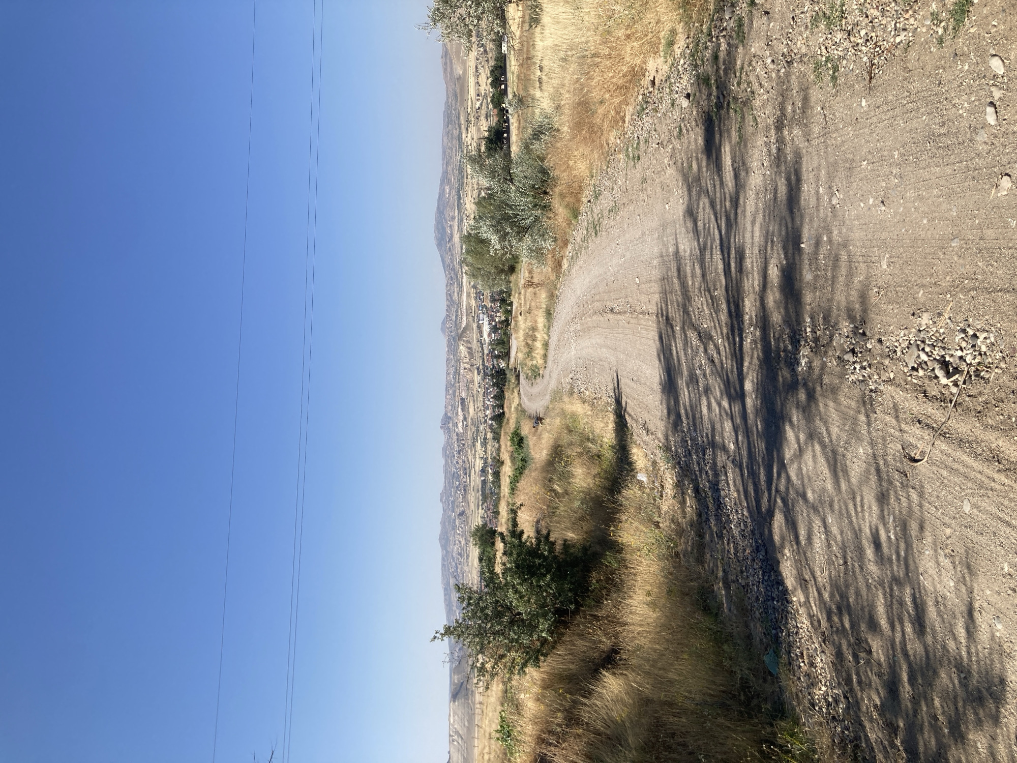 The image depicts a dirt road winding through a dry, grassy landscape under a clear blue sky. In the distance, the city of Avanos İlçe Merkezi, Turkey, is visible with its characteristic rock formations and buildings. The road shows tire tracks and is bordered by dry, yellow grass and scattered rocks. Sparse trees and bushes dot the landscape, casting shadows on the road. Power lines are visible in the upper part of the image, stretching across the sky. The scene appears to be a rural area on the outskirts of the city, bathed in bright sunlight, suggesting a warm, dry climate. There are no people or animals clearly visible in the image.
