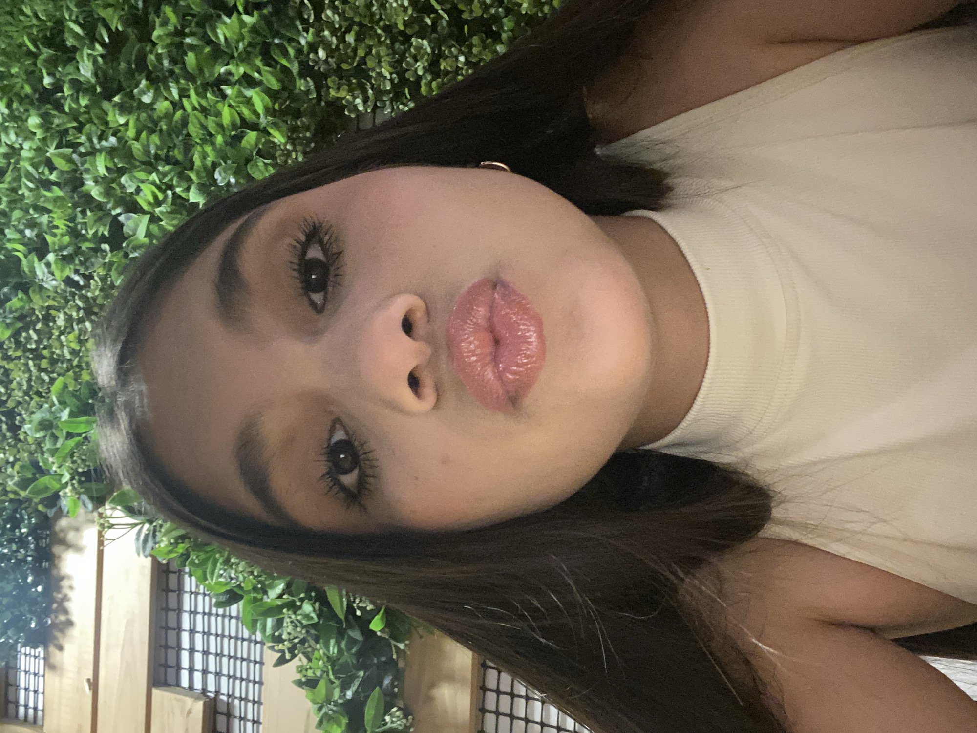A close-up, front-facing portrait depicts a young female individual with dark brown, straight hair parted down the middle. She has brown eyes with visible mascara and defined eyebrows. Her lips are pursed in a 'kissing' gesture, coated with a glossy pink product. She wears a light-colored, possibly off-white or cream, ribbed mock-neck sleeveless top. A small, thin gold hoop earring is visible on her right earlobe. The background behind her left shoulder features horizontal wooden slats intersected by a dark mesh or grate, with green foliage interwoven. To her right, the background consists of a dense wall of green foliage with small leaves and minor white floral elements. The scene is brightly illuminated. This occurs within the city of Pampatar, Venezuela.