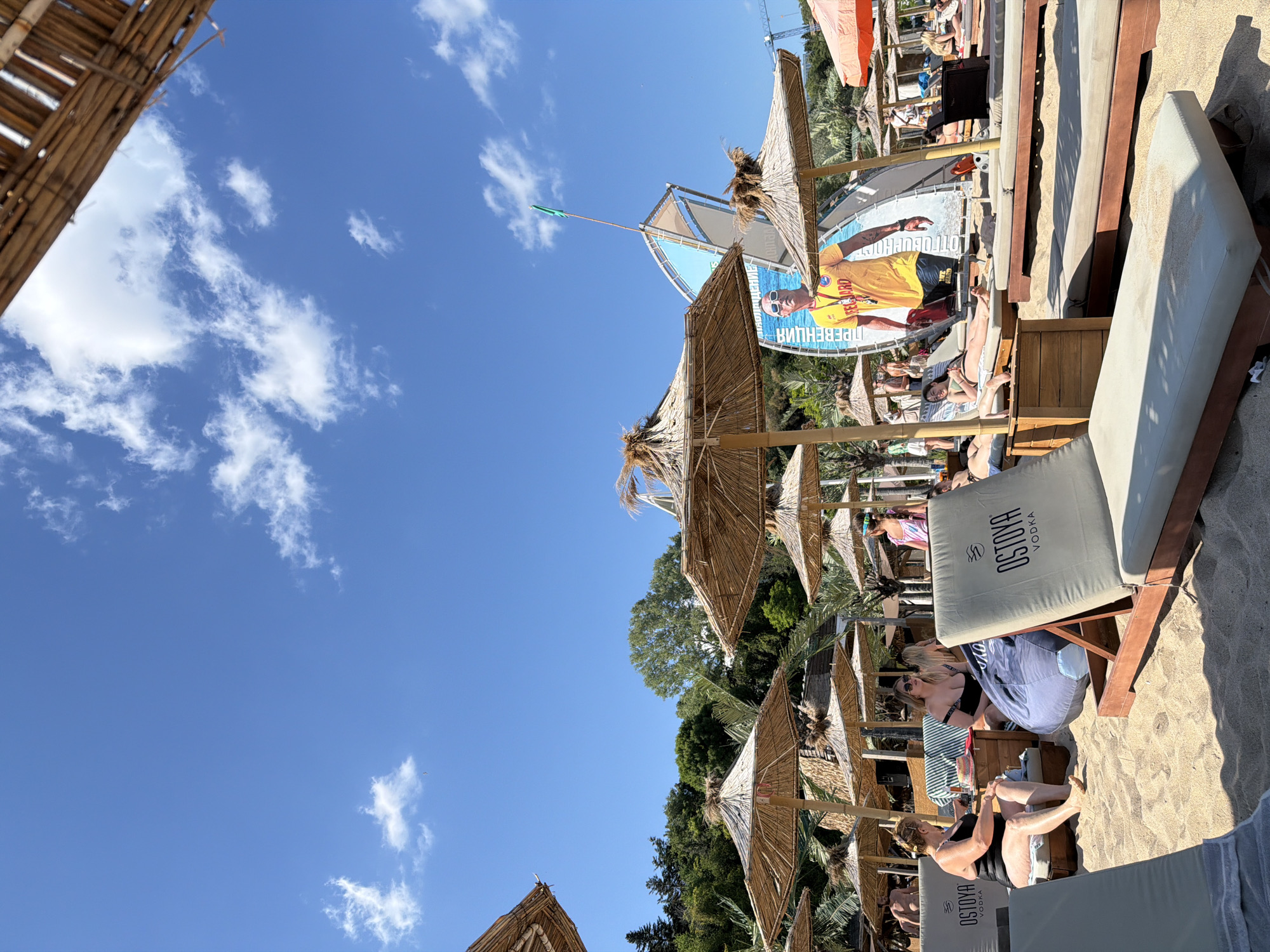 The image depicts a beach scene at Ostoya Beach, a well-known establishment in Varna, Bulgaria. The beach is filled with lounge chairs and straw umbrellas, indicating a recreational setting. In the background, a large billboard features a lifeguard, suggesting safety and leisure activities are promoted. Several people are visible, some relaxing on lounge chairs, others walking or socializing. The overall atmosphere is that of a sunny day at a popular beach resort, with clear blue skies and scattered clouds. The presence of "Ostoya Vodka" branding on the lounge chairs indicates a commercial sponsorship or partnership. The scene suggests a vibrant and active beach environment, typical of summer tourism in Varna.
