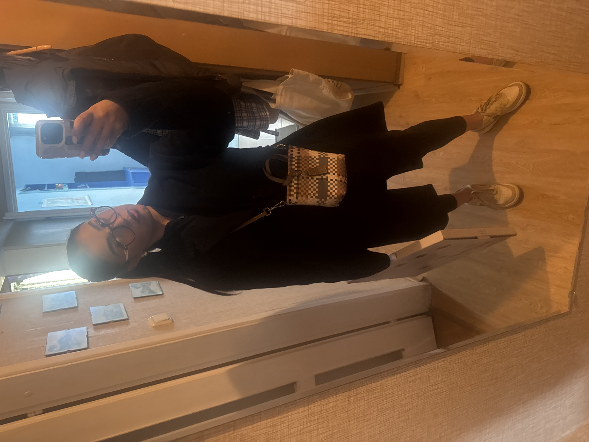 A reflection of a person in a mirror. The person is wearing a black outfit, glasses, and sneakers. They are holding a phone and taking a picture of themselves. The background includes a wooden floor, a window with square panels, and a shelf with some items on it. The location is Unknown, Unknown.