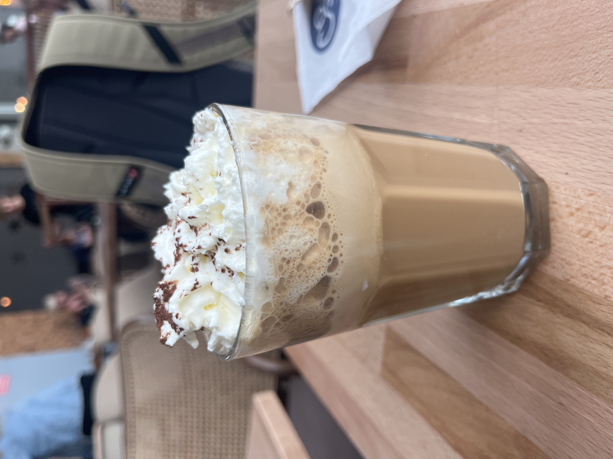 A close-up, eye-level shot captures a tall glass of iced coffee topped with whipped cream and sprinkled with cocoa powder, resting on a light-colored wooden table. The drink is the primary focus, with its frothy layers and creamy topping clearly visible. In the background, slightly out of focus, the interior of a cafe or restaurant is suggested, with patrons seated at tables and a beige jacket draped over a chair. The overall ambiance is relaxed and casual. The photo was taken in Balice, Poland.