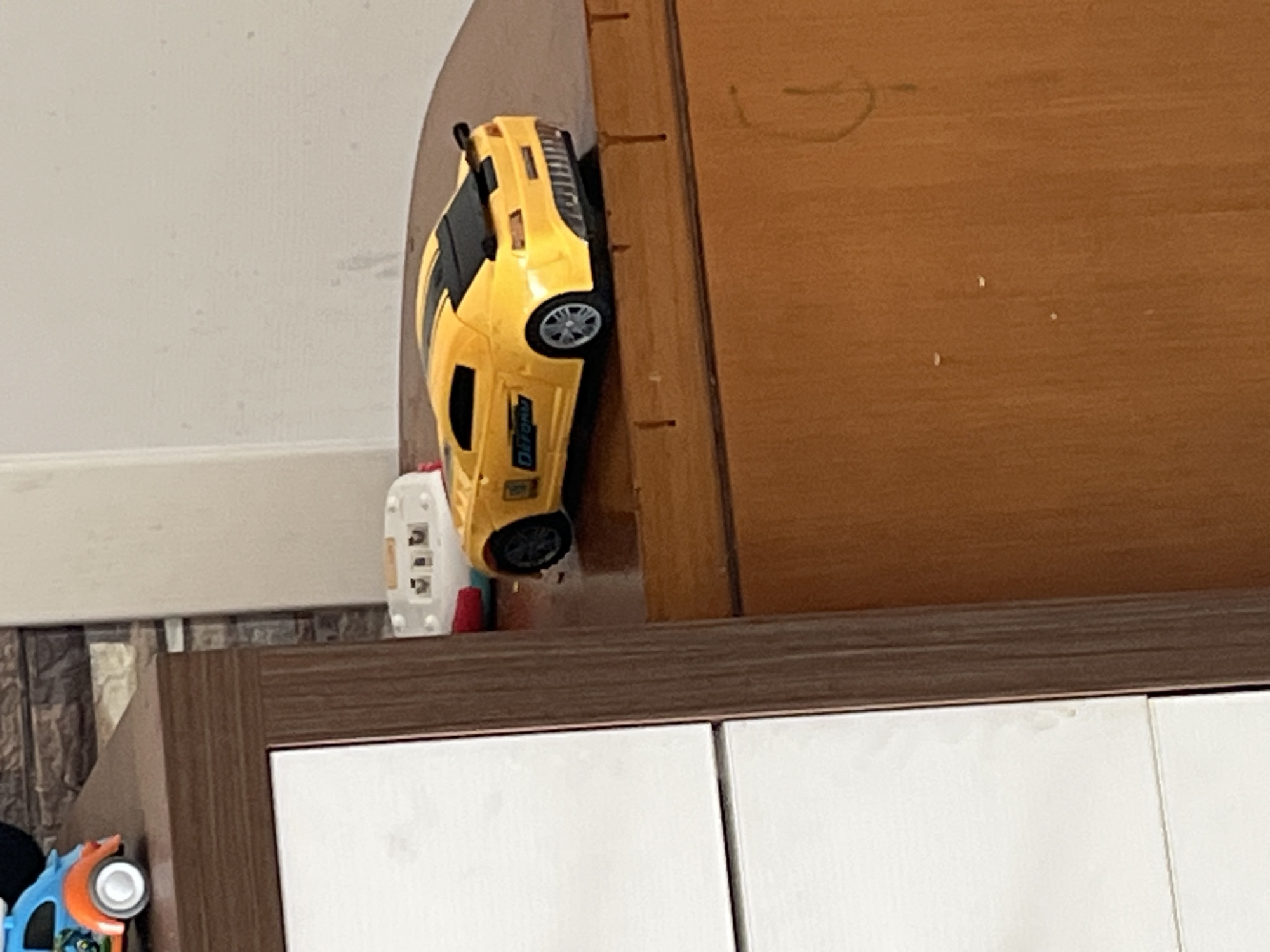 A yellow toy car is precariously balanced on the edge of a wooden surface. The car is a sports car model, with black accents and detailing. It appears to be a die-cast model, possibly a replica of a performance vehicle. The wooden surface is a rich brown color and has a textured grain. Several small, dark marks are visible on the wood, possibly from wear and tear or age. To the left of the car, a small white electronic device with buttons and a screen is partially visible. Further to the left, part of a blue and orange toy vehicle is visible, with a white wheel and orange accents. The background on the left is a textured wall, likely in a home setting. The overall impression is of a child's play area or a shelf where toys are stored. There is no discernible activity taking place, as the car is stationary. The lighting suggests an indoor environment, possibly during the daytime. There is no visible text on the toy car that can be read clearly.