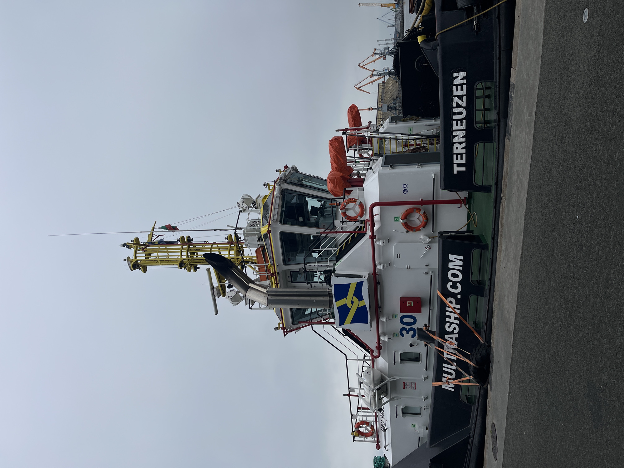 The image shows a tugboat named "TERNEUZEN" docked at a pier. The tugboat is predominantly white with black accents. "MULTRASHIP.COM" is printed on its side. There is a flag with a blue and yellow chain emblem flying. The number "30" is visible on the hull. In the background, cranes and industrial structures are visible, typical of a port setting. Based on the name on the tugboat "TERNEUZEN" it is not located in the city of Burgas.