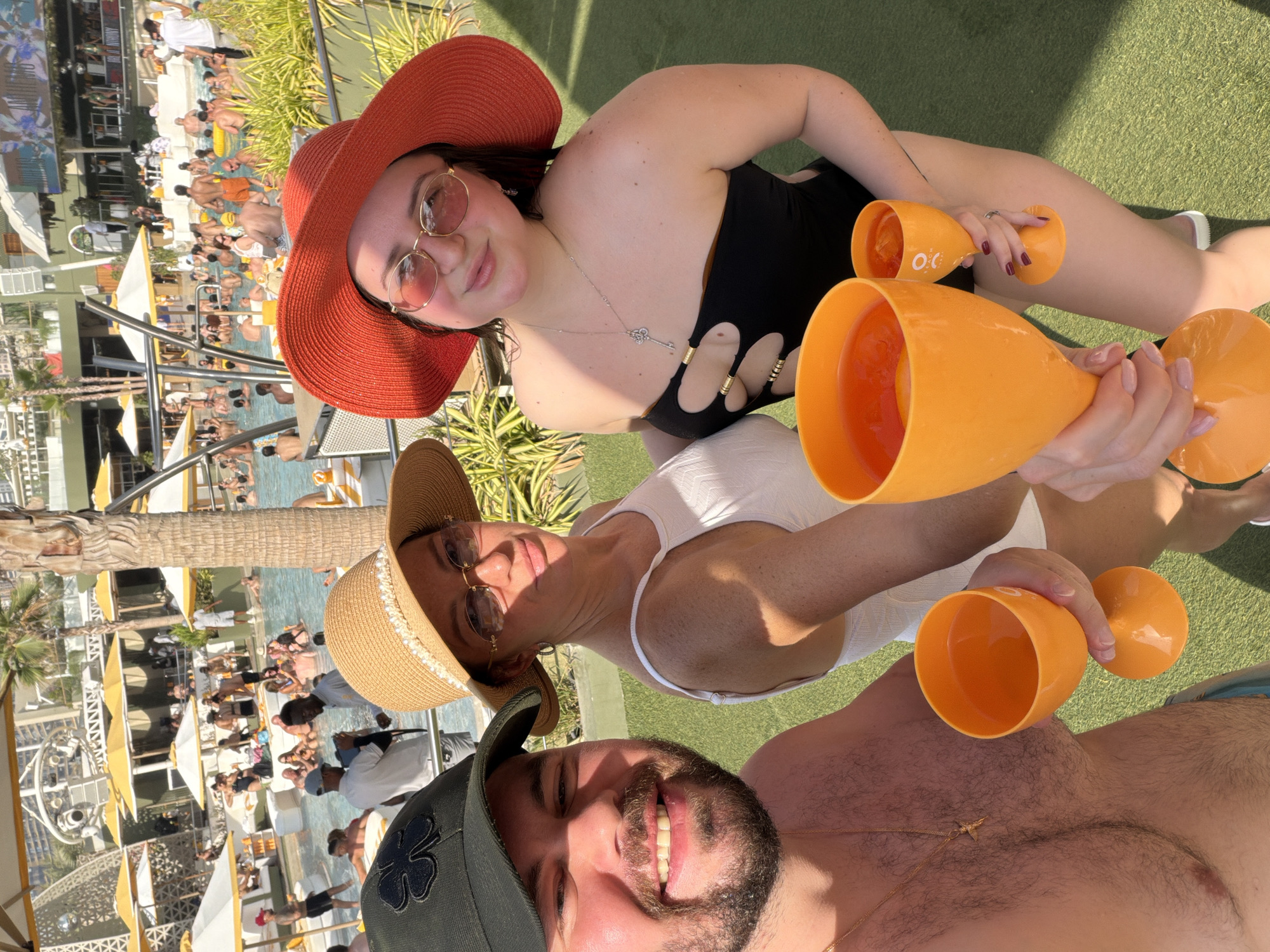 In a Dubai resort, three individuals pose for a selfie by a pool. The man on the left, wearing a green baseball cap and a gold chain, smiles at the camera, holding an orange glass of drink. Next to him, a woman in a wide-brimmed straw hat and white top also looks towards the camera, with an orange drink in her hand. On the right, another woman, sporting a large red hat and sunglasses, wears a black cut-out swimsuit and holds a similar orange drink. The background reveals a bustling pool area with numerous people, yellow umbrellas, and palm trees, suggesting a vibrant resort atmosphere.