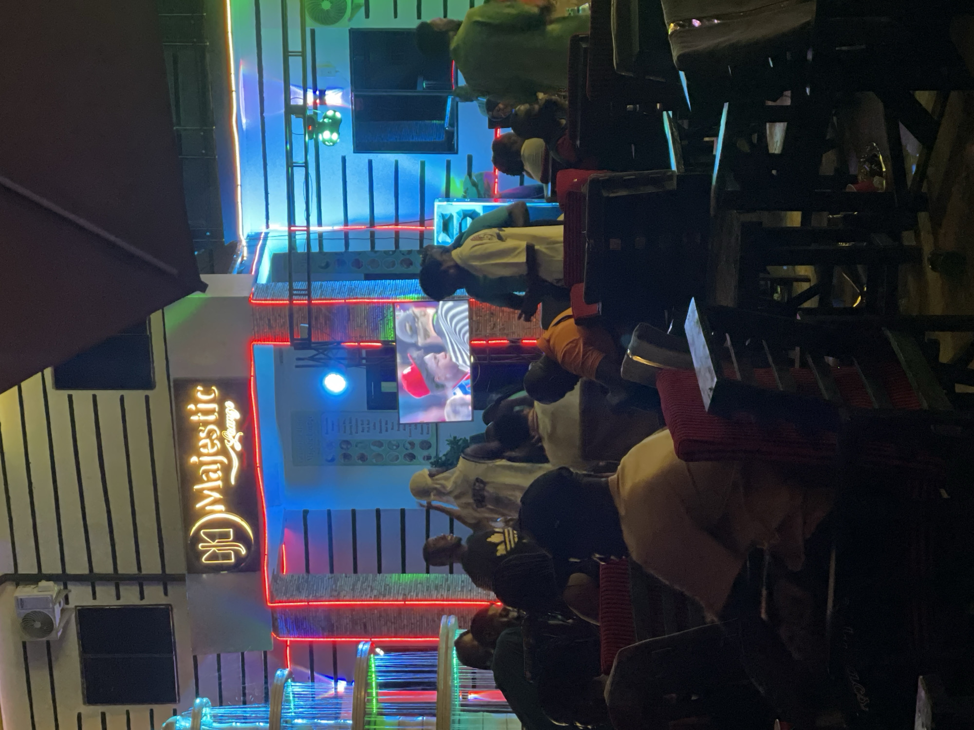 The image depicts an outdoor scene at night in Tumfure, Nigeria. People are seated on high stools, possibly at a bar or lounge. The "Majestic Lounge" sign is visible on the side of a building, illuminated in gold. The building has vertical stripes and neon lights, including blue, red, and green colors. A television screen displays content. Some speakers or audio equipment are installed above the seating area. The environment is dimly lit overall, and many details are obscured by darkness.