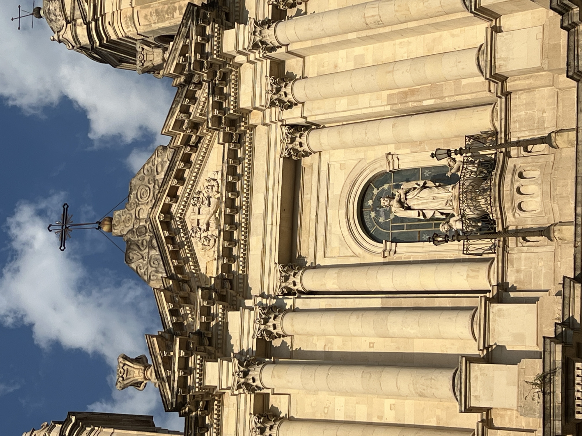 This is a vertical, eye-level, close-up shot of the facade of a grand, ornate building, likely a church, in Catania, Italy. The architecture is Baroque, characterized by elaborate carvings, columns, and decorative elements. The stone material of the building appears to be light-colored, possibly sandstone or limestone, and is illuminated by bright sunlight, casting distinct shadows.
The primary subject is the architectural detail of the building. There are several large, fluted columns adorning the facade, topped with intricate Corinthian capitals. Between the columns are carved friezes and pediments, featuring figurative sculptures and geometric patterns. In one prominent section, a semicircular arched window is visible, containing a stained-glass depiction of what appears to be a religious scene with figures. A statue of a robed figure, possibly a saint, is positioned on a small balcony above this window, accompanied by another smaller figure. A wrought-iron railing and a decorative lamppost are also present on this balcony.
At the very top of the visible structure, a cross with a distinctive pointed finial stands against a backdrop of a bright blue sky dotted with fluffy white clouds. This suggests it is daytime, likely late morning or early afternoon, given the strong sunlight and clear skies. The clouds indicate fair weather. The overall impression is one of grandeur, history, and religious significance. No text is visible in the image.