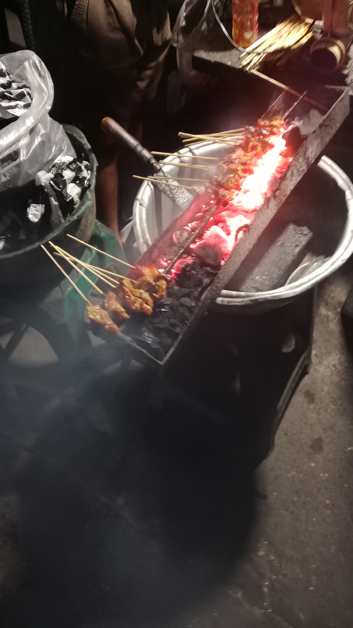This is a medium-shot, eye-level, vertical-orientation photograph taken at night on a street in Denpasar, Indonesia, capturing a scene of satay being grilled. In the foreground, several skewers of meat, appearing to be chicken satay, are positioned over glowing red charcoal embers in a long, rectangular grill. The satay is partially cooked and some smoke is visible rising from the grill. To the left of the grill, a large, black plastic bag filled with what appears to be white and black packaging material is seen, next to a dark, cylindrical object that could be a bucket or a container. Behind the grill, a portion of a person's legs, clad in dark trousers, is visible, suggesting the presence of a vendor or customer. In the background, out of focus, there are more indistinct objects, including what might be additional food preparation materials and possibly a bottle. The overall ambiance is that of an outdoor food stall or street vendor at night, common in the bustling urban environment of Denpasar.