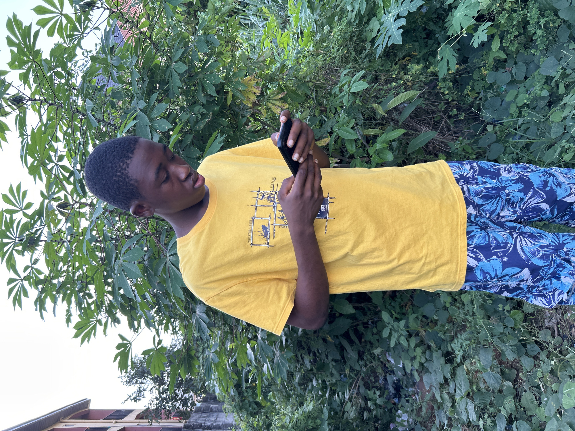 A young man is outdoors in Ihiagwa, Nigeria. He is standing near a variety of leafy plants and trees with cassava leaves, holding a dark-colored phone in his hands, looking at it. He is wearing a yellow t-shirt and blue floral-patterned pants. Part of a building is visible in the background.