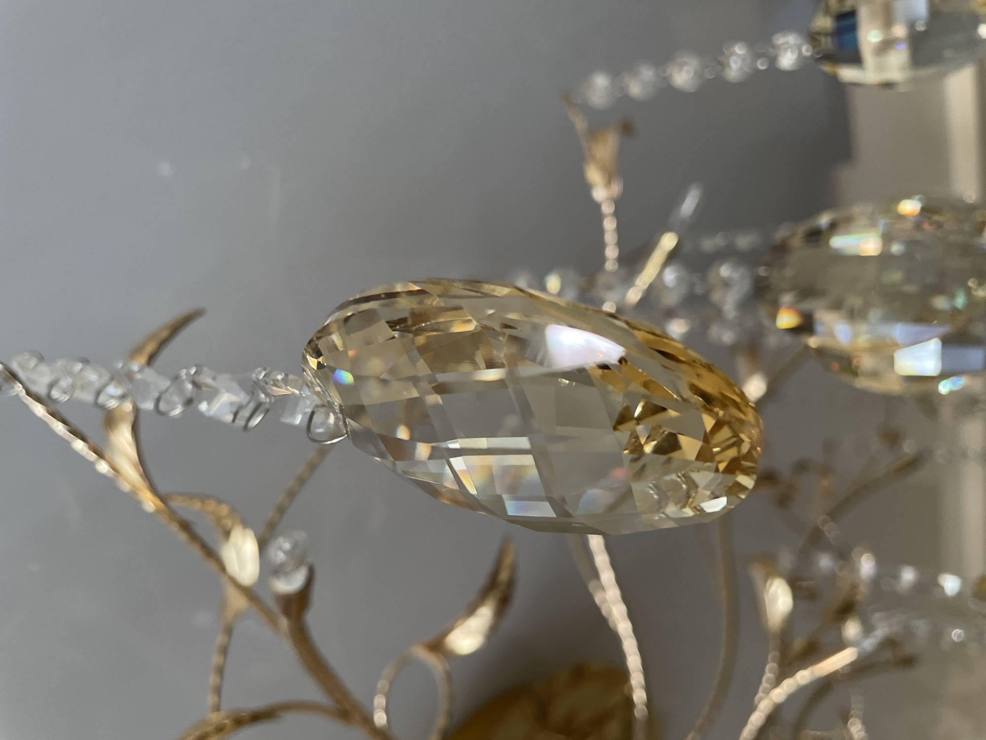 Close-up view of a decorative chandelier with multifaceted golden and clear crystal components, and small white beads hanging from gold-colored, leaf-shaped fixtures. The background is a blurred neutral gray wall. Location cannot be determined from the image and is unlikely to be Izhevsk, Russia.