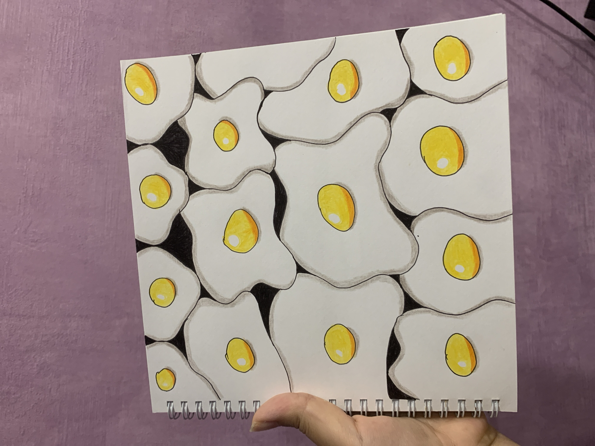 The image shows a notebook with a drawing of fried eggs on its cover. The eggs are arranged in a repeating pattern across the cover. The notebook is held by a person's hand, and it is placed on a purple textured surface. The drawing is done with black marker and colored pencils, with yellow yolks and white egg whites. There are shadows drawn around the eggs to give them a three-dimensional appearance. The location of Asha, Russia, is not depicted in this image.