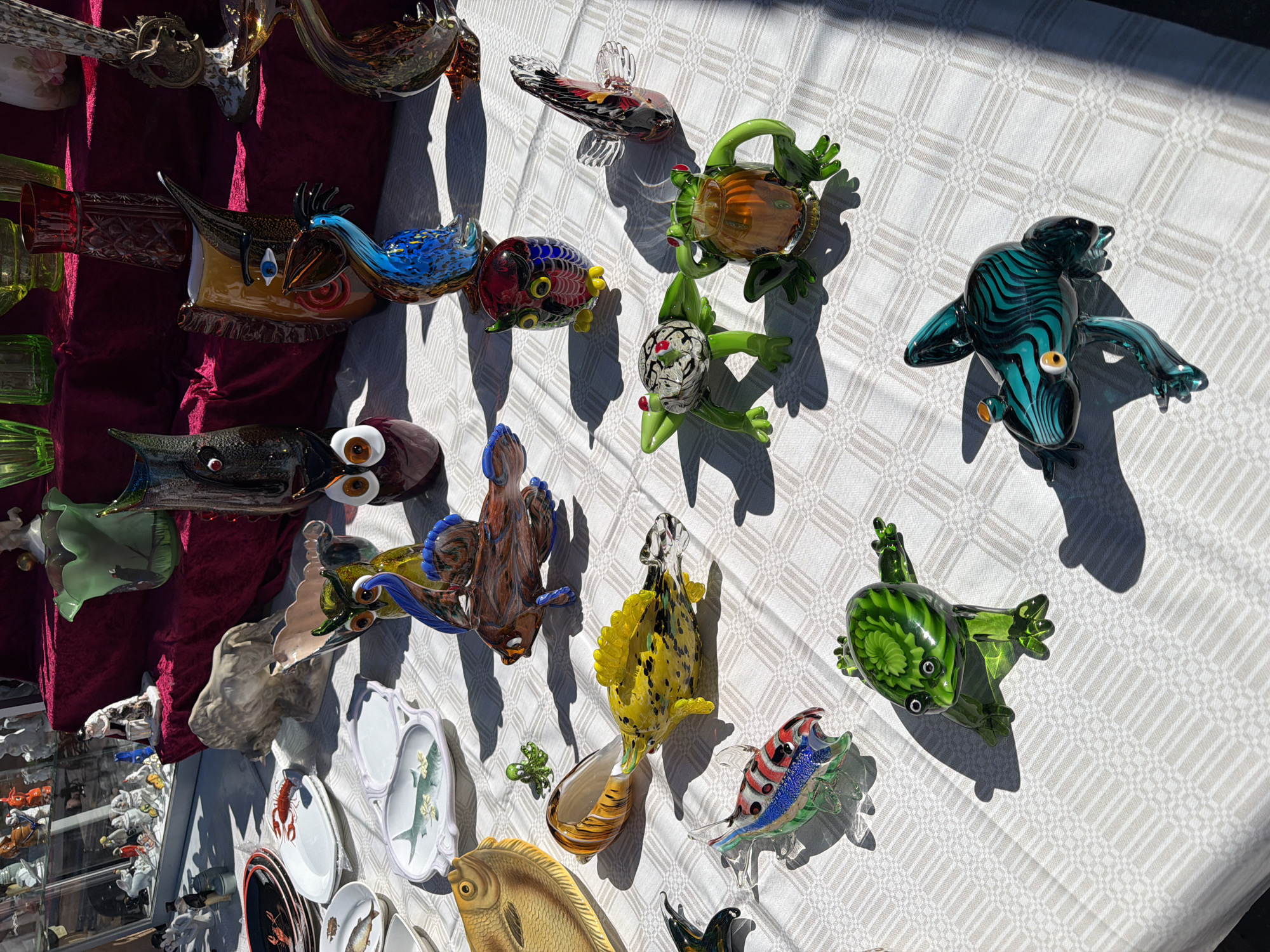This image captures a brightly lit outdoor market stall in Riga, Latvia, showcasing an array of glass and ceramic items. The foreground features a table draped with a white tablecloth bearing a subtle checkered pattern. On this table, numerous small, colorful glass figurines are arranged, including several frogs (green, blue-and-black striped, and white with red spots), various fish (yellow speckled, multi-colored striped, and a larger brown-and-blue stylized fish), a small green octopus, and multiple stylized glass birds (including an owl-like figure and a blue pelican-like bird). To the left, several ceramic plates are visible: two black plates with red crayfish designs, two white rectangular plates featuring painted fish and crayfish, and a large, textured yellow ceramic platter shaped like a fish.
Behind this display, a second table is covered with a deep red fabric, presenting larger and more intricate glass art. These include tall, dark glass sculptures resembling owls or abstract faces, one with a distinct blue eye, and a prominent blue and brown glass sculpture resembling a bird. Additional glass vases and pitchers in shades of green, red, and amber are also present. In the upper left background, behind the red-draped table, a glass-encased display contains numerous white ceramic or porcelain figurines depicting various forms and figures. Strong, defined shadows across the tablecloth indicate direct overhead sunlight. No individuals are visible in the scene.