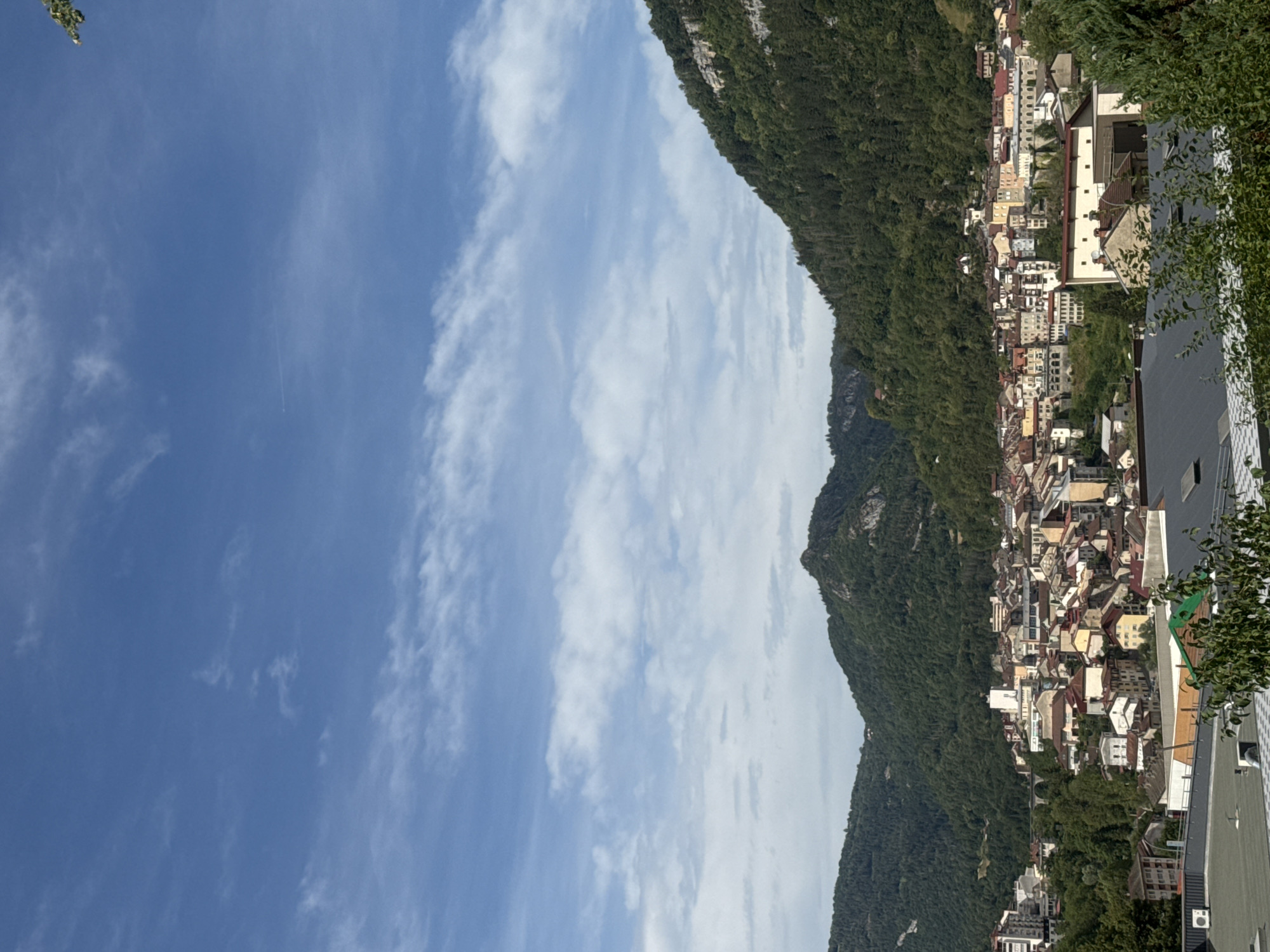 This image captures a panoramic view of the city of Saint-Claude, France, nestled within a lush, mountainous landscape. The sky is a vibrant blue, adorned with wispy white clouds, suggesting a clear and pleasant day. The foreground is dominated by the cityscape, a dense collection of buildings with red and beige tiled roofs, interspersed with trees. The architecture appears varied, with some taller, more modern structures standing out amongst older, more traditional buildings. The city extends up the slopes of the surrounding mountains, which are covered in dense green forests, creating a dramatic backdrop. One prominent mountain peak, partially shrouded in shadows, juts out in the middle distance, while another, larger forested mountain range forms the right side of the frame, its steep slopes dotted with rocky outcrops. In the distance, a viaduct or bridge can be seen spanning across a valley within the city. The overall scene conveys a sense of a charming town integrated harmoniously with its natural surroundings.