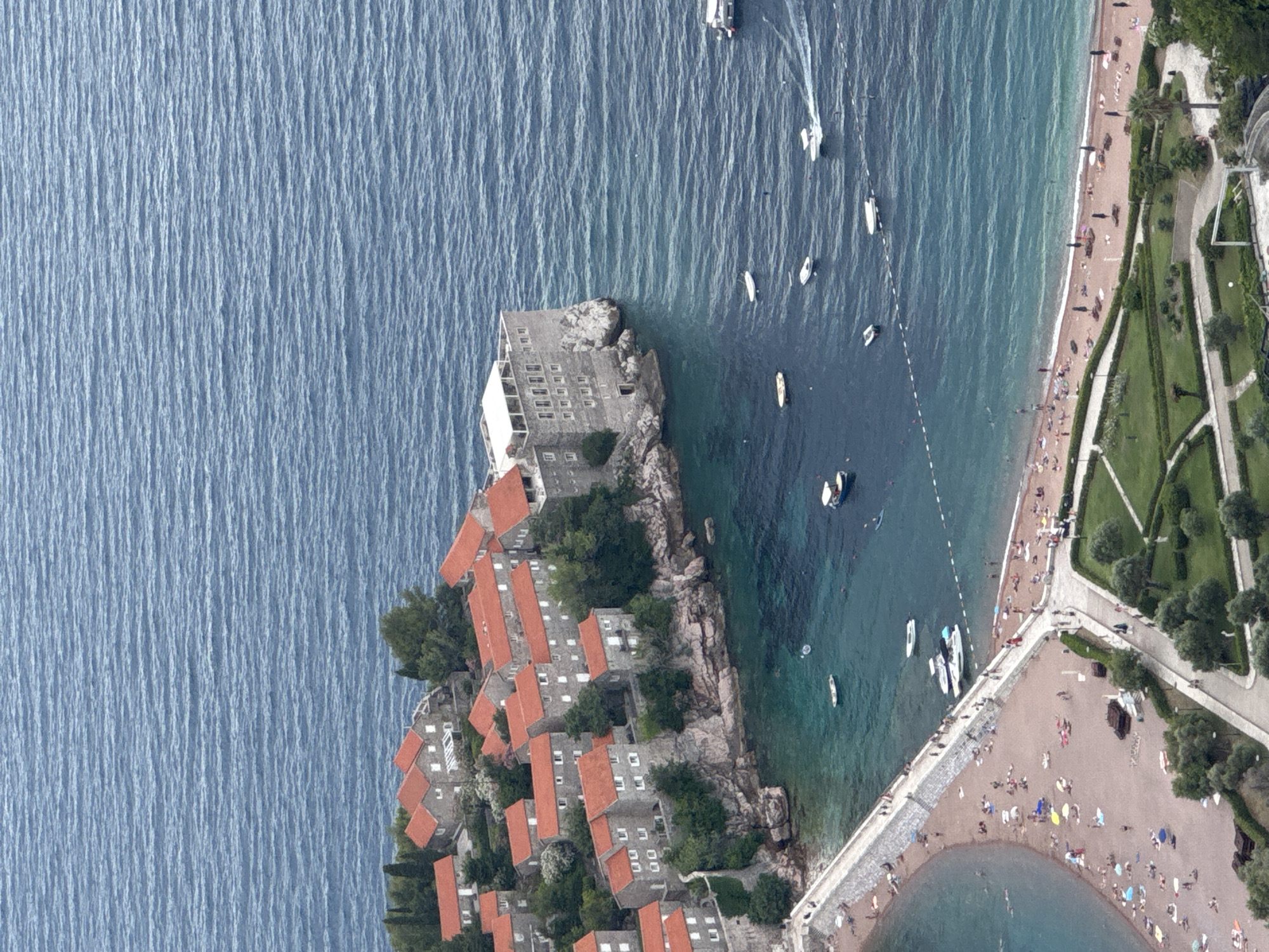 This aerial shot captures the picturesque Sveti Stefan island in Montenegro. The island, connected to the mainland by a narrow causeway, is dotted with red-roofed stone buildings that appear to be historic residences or hotels. The surrounding waters are a deep blue, with gentle waves creating a textured surface.
Several boats are scattered across the sea, including a few speedboats leaving white wakes, and smaller vessels like kayaks or paddleboards. On the right side of the image, a sandy beach is bustling with activity. Numerous people are sunbathing, walking along the shore, and swimming in the clear turquoise water. Umbrellas are set up, providing shade for beachgoers.
Adjacent to the beach is a meticulously landscaped park with green lawns, trees, and winding pathways. The architecture of the island and the presence of lush greenery suggest a warm, sunny day, likely during the tourist season. The scene evokes a sense of leisure and holiday enjoyment. The time of day appears to be daytime, given the sunlight casting shadows and illuminating the water. No visible text is present in the image.