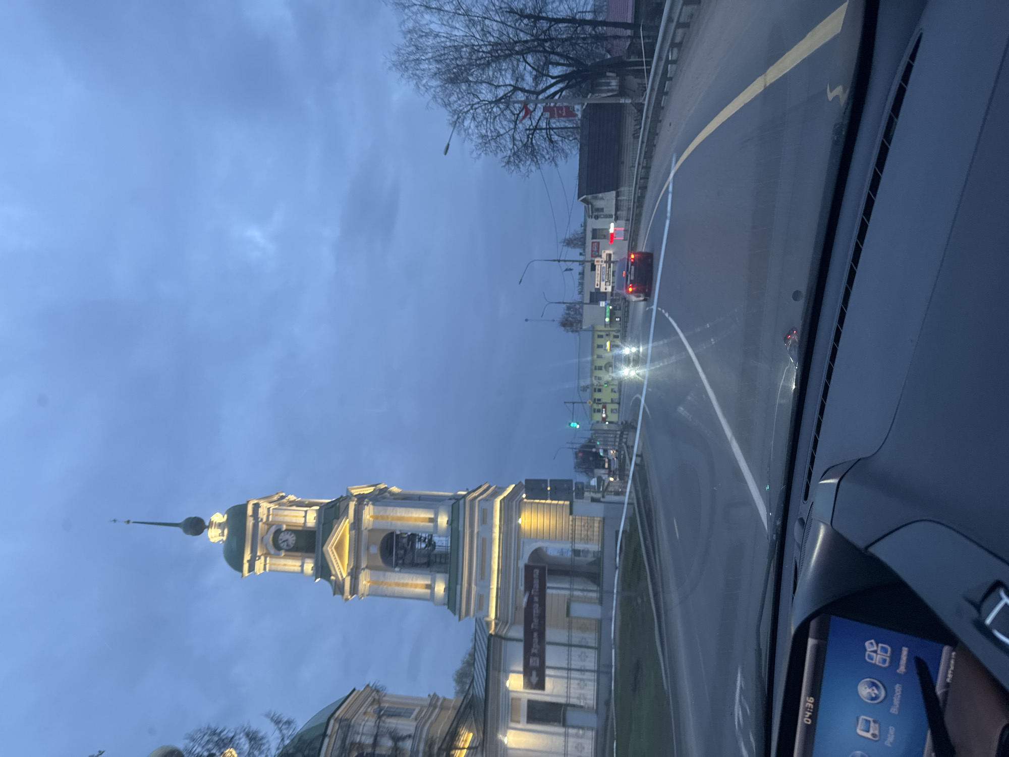 The image, taken from inside a car, shows a view of a road and a church in the city of Ново-Никольское, Russia. The road, marked with yellow and white lines, stretches ahead, with cars visible in the distance. To the left of the road, a cream-colored building with a green dome and a tall, slender bell tower with a clock on its face stands prominently. The sky above is a muted blue-gray. The car's dashboard is visible in the foreground, showing a digital clock displaying "04:36".