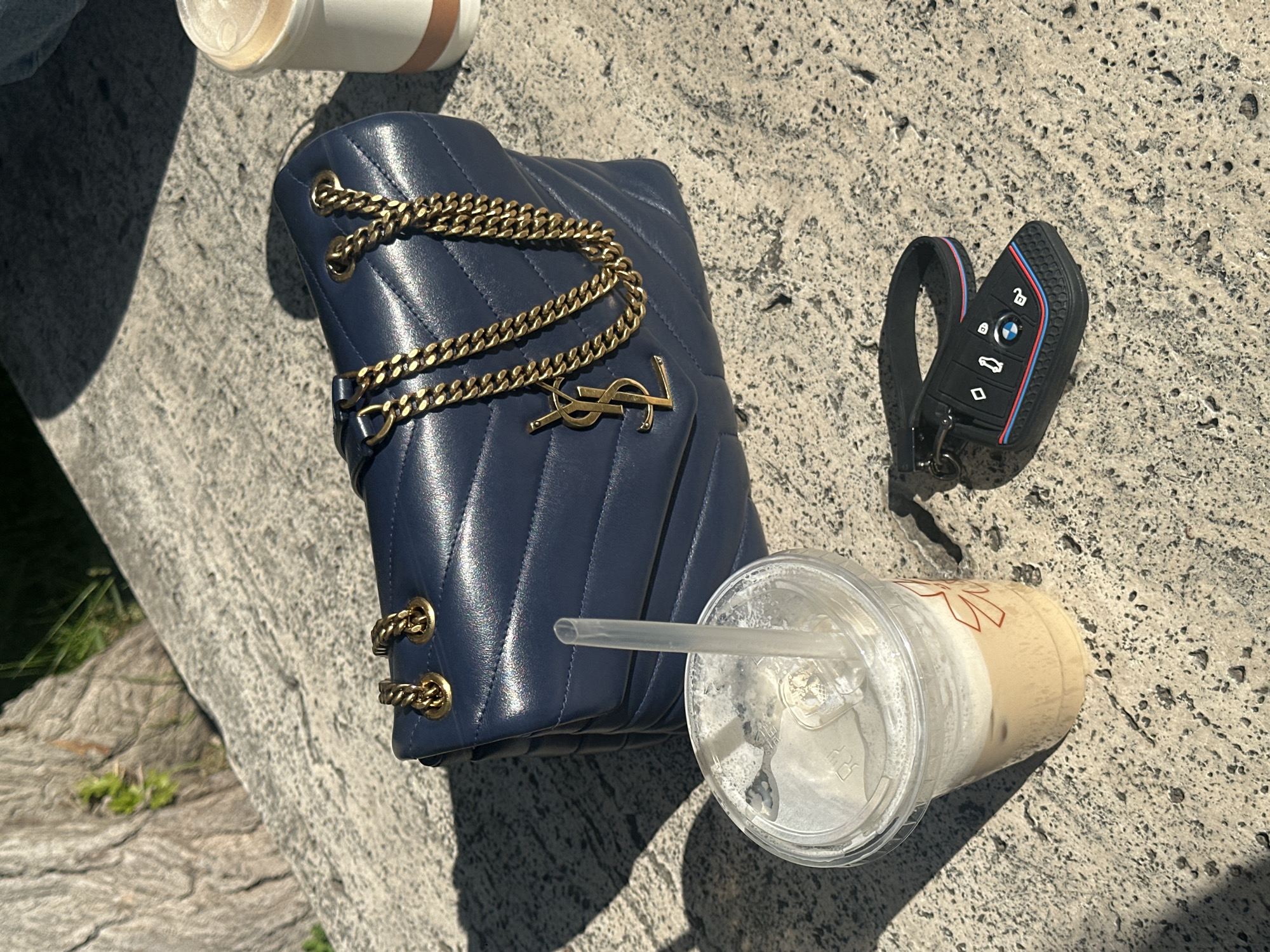 The image shows a light grey stone surface, likely a table or bench, with a blue Yves Saint Laurent quilted leather purse with a gold chain strap and YSL logo, a black BMW car key with a colorful M stripe accent, and two partially consumed beverages in disposable cups. One cup is clear and contains a drink with ice and a straw, and the other appears to be a coffee in a white cup with brown accents. The image appears to be taken outdoors in daylight. Given the architectural style of Yerevan, Armenia, it is possible that this scene is located in one of the city's outdoor cafes or parks. No people are visible in the image.