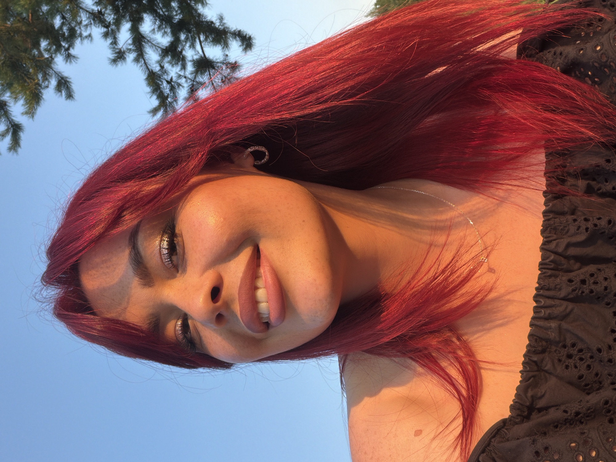 A young woman with bright red hair and a black off-the-shoulder top smiles gently at the camera. She wears subtle makeup, including shimmering eyeshadow, and a thin silver necklace with a small pendant. A circular earring glints in her right ear. Her skin is dusted with freckles, particularly on her left shoulder. The background is a clear blue sky, with the tips of pine trees visible on the right. The golden hour lighting casts a warm glow on her face and hair. The image appears to have been taken outdoors, possibly in a park or natural area within Tolyatti, Russia, during the late afternoon or early evening.