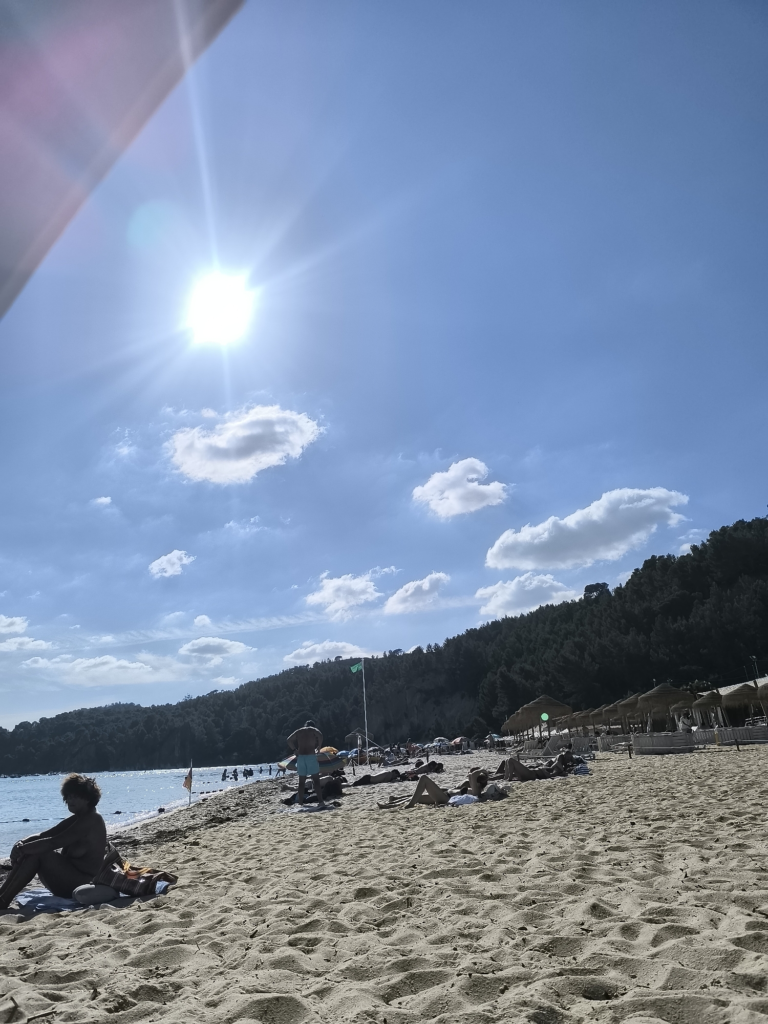 This is an eye-level, wide-angle shot of a beach scene on a sunny day. The sky is a clear blue with some scattered white clouds. The sun is high in the sky, creating a bright glare in the upper left quadrant of the image. The beach is covered in light-colored sand, with footprints visible in the foreground.
In the mid-ground, several people are visible. On the left, a woman is sitting on a towel, facing away from the camera, with her legs crossed. She appears to be wearing a bikini top. Further down the beach, a man in blue shorts stands with his back to the camera, looking out towards the water. Several other people are lying on towels or lounging on the sand, some in swimwear, others partially or fully clothed. In the water, several figures are visible, some swimming or wading.
The background features a body of water that appears calm. Along the shoreline, there are several straw-covered umbrellas and lounge chairs, suggesting a resort or organized beach area. A line of buoys extends into the water. Behind the beach and resort area is a densely wooded hillside. A green flag is visible on a pole on the hillside, near the beach. The overall atmosphere is one of leisure and recreation.
While the image depicts a beach scene, it is not possible to definitively identify the specific location within Setúbal, Portugal without additional information.