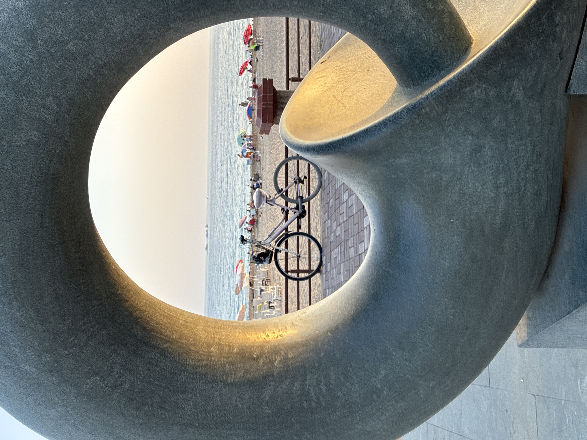 In Alanya, Turkey, a bicycle is parked on a paved walkway overlooking a crowded beach. The scene is framed by a large, abstract stone sculpture. The beach is dotted with numerous colorful umbrellas and lounge chairs, with many people visible sunbathing and enjoying the water. The ocean stretches to the horizon under a hazy, light-colored sky. A few boats are visible on the water in the distance. The sculpture, made of a coarse, grey stone, has a curved, organic form with sunlight glinting off its polished inner surface. The walkway is paved with rectangular stone tiles, and a wooden railing separates it from the beach.