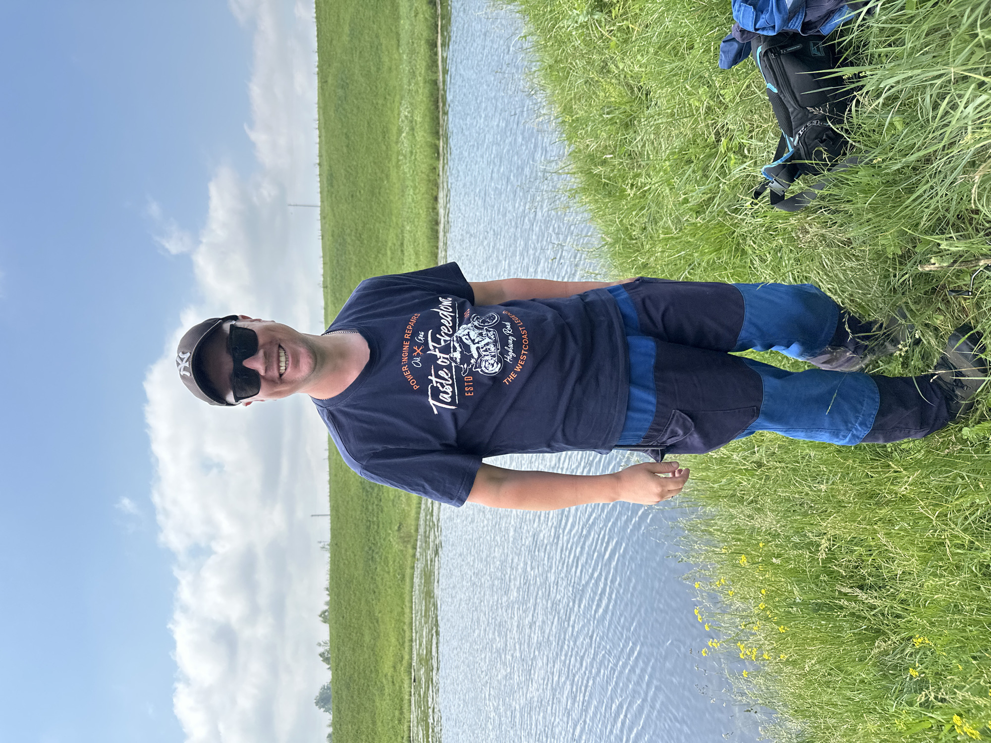 A man is standing on the grassy bank of a body of water, likely a river, with a green embankment behind him under a partly cloudy sky. He is wearing a dark blue t-shirt with a graphic print that reads "Taste of Freedom" and features a motorcycle, sunglasses, and a dark baseball cap. His pants are a combination of dark blue and lighter blue panels. To his right, on the grass, are a black fanny pack and a blue backpack. The overall setting appears to be a rural or park area, possibly in the village of Sredneye Kechevo, Russia, given the description.