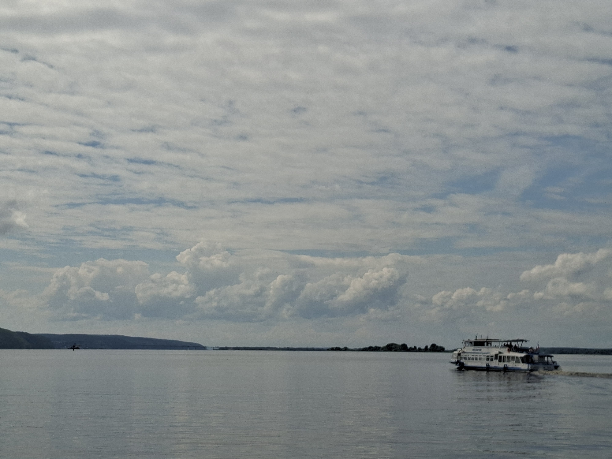The media file depicts a wide, calm body of water, likely a river, under a cloudy sky. The water is a muted gray-blue, reflecting the overcast conditions. In the distance, a dark, undulating treeline forms the horizon, punctuated by a bridge on the right and a large hill or mountain on the left.
The main subject of the image is a white passenger boat moving from right to left across the frame. The boat has multiple decks and appears to be carrying passengers, though they are too small to discern details. The boat is leaving a visible wake, indicating its movement through the water.
The sky is filled with a mix of gray and white clouds, with patches of blue sky visible in between. The clouds are varied in texture, with some appearing wispy and others more bulbous and defined. The lighting suggests it is daytime, likely afternoon, given the diffused light.
There are no explicit cues to suggest the specific location beyond the provided context of Kazan, Russia. The scene evokes a sense of peaceful travel or a serene day on the water. No visible text is present in the image.
