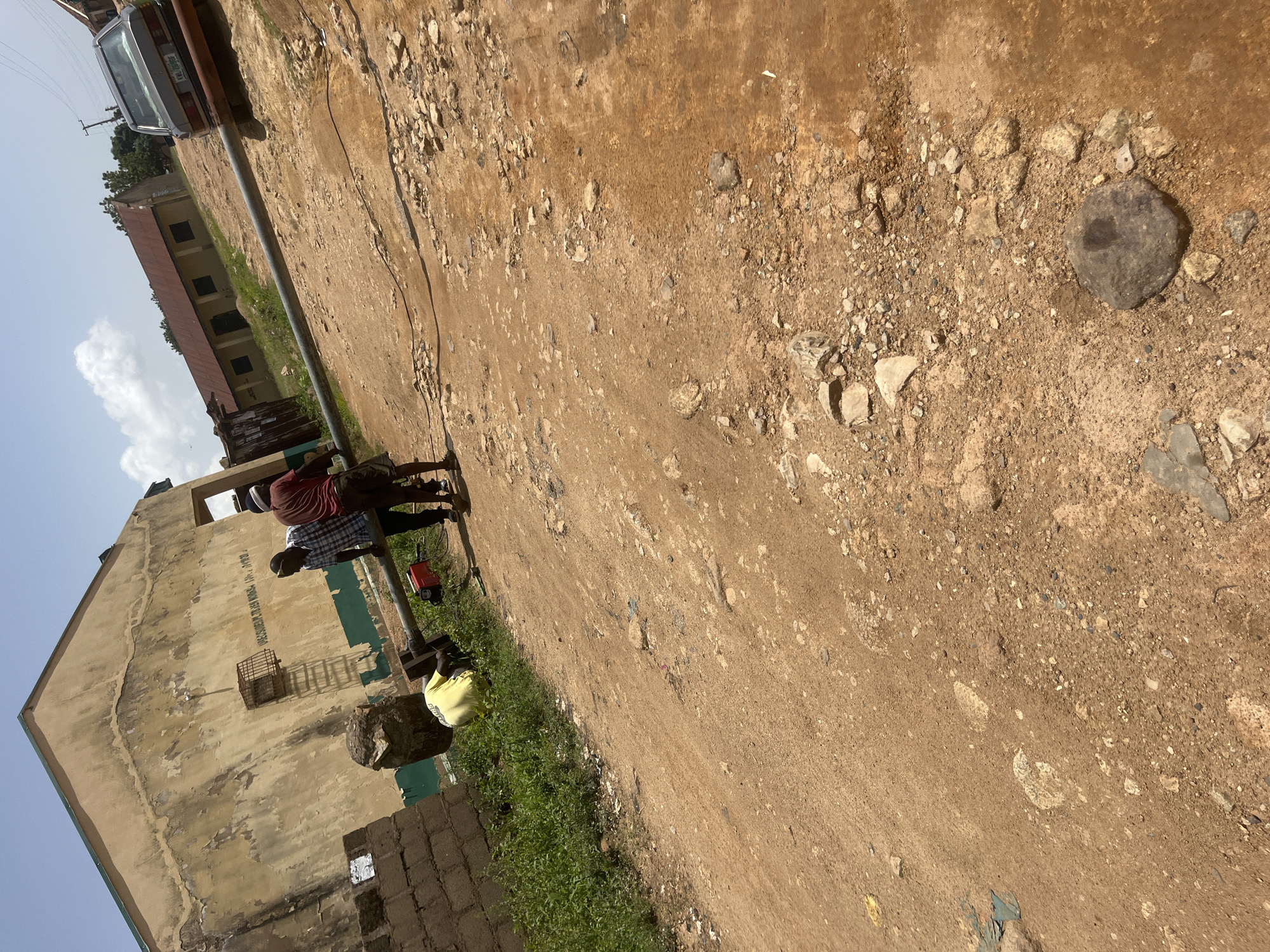 The media file depicts an outdoor scene with a building and a dirt road, likely in Gura Top, Nigeria. Three individuals are present. One person, wearing a red and white striped shirt, stands near the building. Another person, in a blue and white checkered shirt, stands next to them. A third person, wearing a yellow shirt, is sitting down further down the road.
The building appears to be weathered, with peeling paint on its beige exterior. Green trim runs along the bottom of the building, and there are several windows visible. A metal cage is attached to the wall of the building. Part of a brick wall is visible in the foreground. A car is parked on the left side of the image, further down the road. The ground is a mixture of dirt, gravel, and scattered rocks. Some green vegetation is growing along the edge of the road next to the building. A red object, possibly a tool or container, is on the ground near the people. The sky is bright, suggesting it is daytime. The overall impression is of a rural or semi-urban environment, possibly during fair weather. The individuals appear to be engaged in casual activity or taking a break.