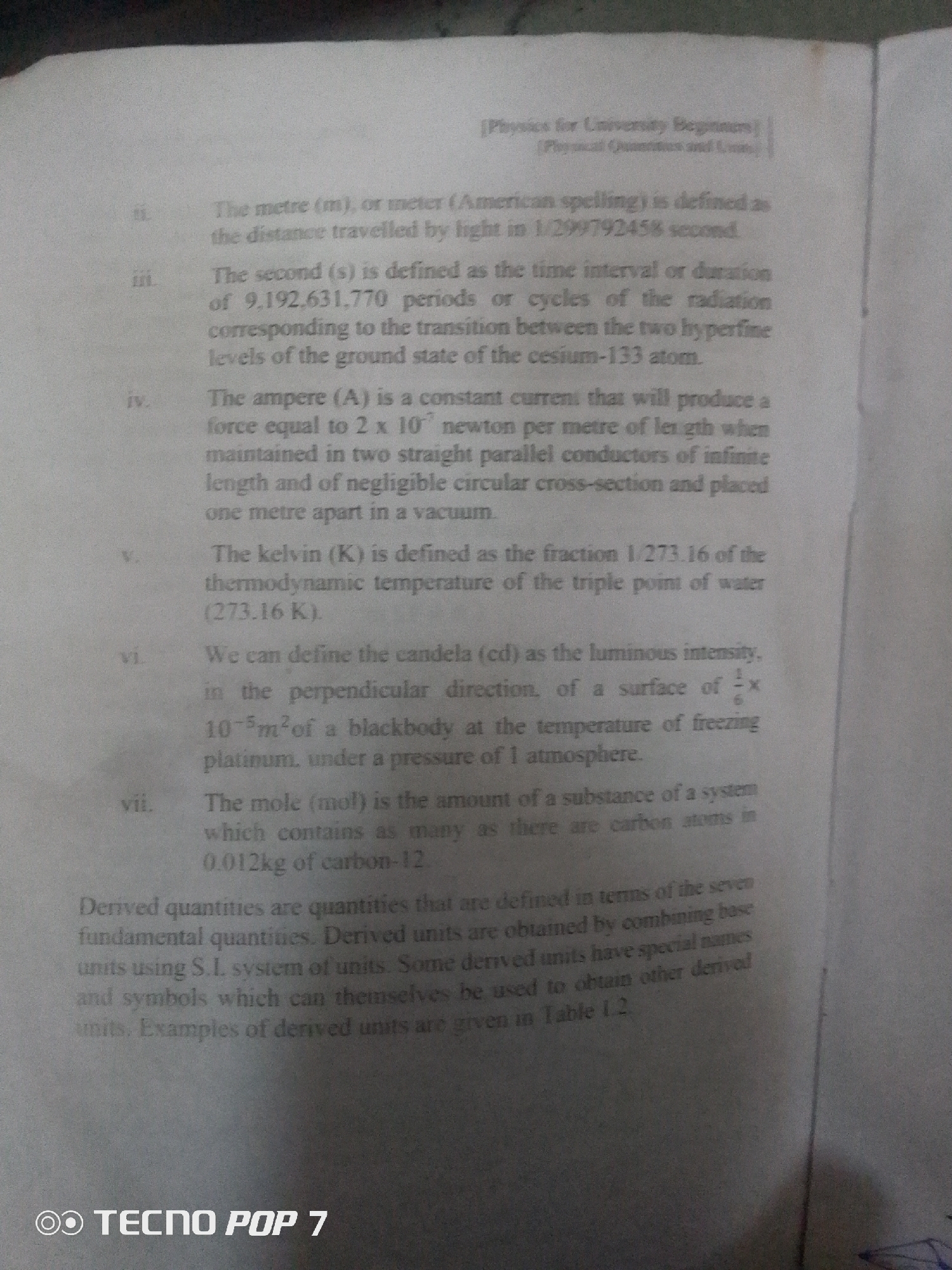 This is a close-up, overhead shot of an open book lying flat, likely on a table. The page displays text from a physics textbook. The subjects are the text itself, with the primary object being the book and paper. The scene is a generic indoor setting, likely a study area or classroom.
The activity taking place is the presentation of educational content, specifically definitions of physical units like the meter, second, ampere, kelvin, candela, and mole. The text also defines derived quantities and units.
Notable details include the clear, black text on a slightly off-white page. The lighting appears to be artificial, given the uniform brightness and lack of shadows suggesting natural light. There are no people visible. The text contains mathematical formulas and numerical values, indicating a scientific or technical subject. A small, possibly smudged, blue ink mark is visible in the bottom right corner. At the very bottom of the image, a logo for "TECNO POP 7" is displayed, indicating the device used to capture the image. The text is in English.