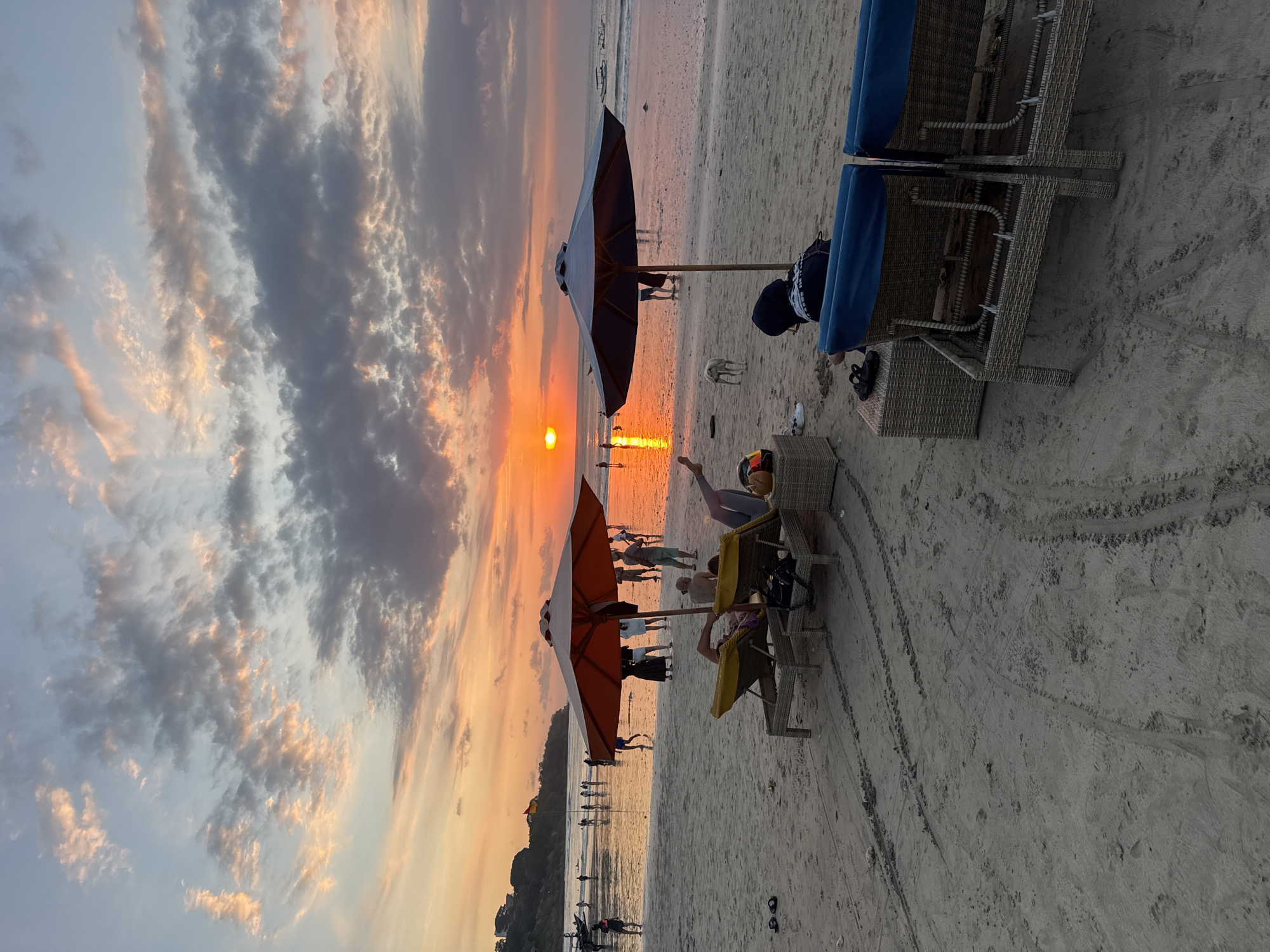 This image captures a vibrant sunset scene on a sandy beach in Kuta Selatan, Indonesia. The time of day is clearly evening, with the sun positioned low on the horizon, casting a brilliant orange and red glow across the sky and reflecting on the water. Scattered clouds, some dark and others illuminated with warm sunset colors, are visible overhead.
In the foreground and mid-ground, the beach is furnished with amenities for relaxation. Two large beach umbrellas stand out: one dark blue/black on the left and another bright orange/red towards the center-right. Several wicker-style beach chairs and loungers are present, some with blue cushions and one with a yellow cushion. A person wearing a blue hoodie is seated on a chair in the right foreground, while another individual is seen relaxing on a yellow-cushioned lounger. A small, light-colored dog stands near one of the umbrellas.
Numerous other people are scattered across the beach, some walking along the shoreline, others standing or gathered in groups, largely silhouetted against the bright light of the setting sun. Their activity suggests they are enjoying the beautiful evening atmosphere. The sand shows various footprints and what appear to be tire tracks from beach vehicles. The ocean is calm, with the sun's reflection creating a shimmering path on the water's surface. The overall mood is peaceful and leisurely, characteristic of a tropical sunset. No discernible text is visible in the image.