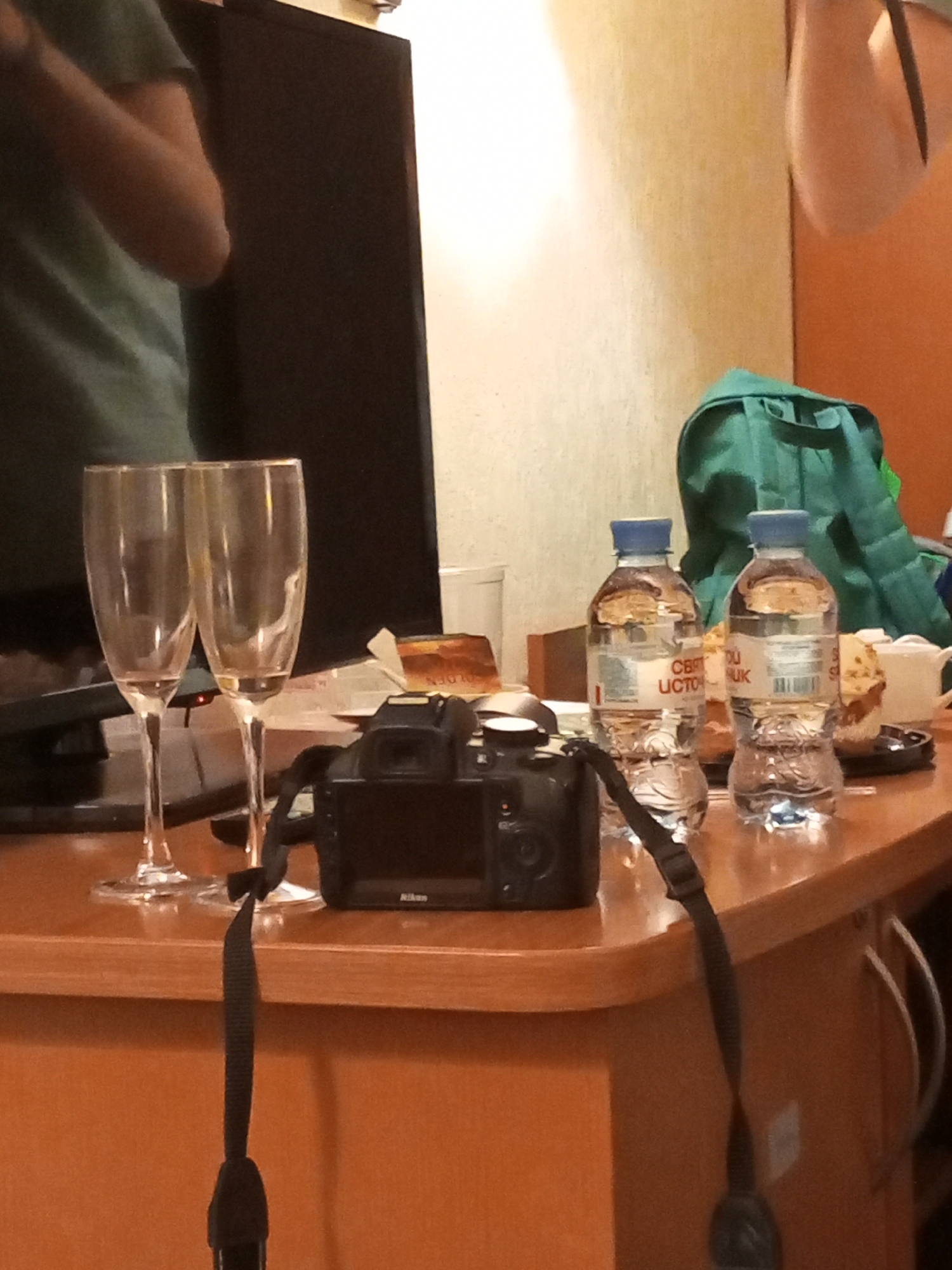 The image shows a room setting with two champagne flutes, a black Nikon camera with a strap, two bottles of water labeled "Cebu Источник" (Cebu Source), a green bag, and partial views of individuals. The setting implies a possible celebration or gathering. The presence of "Cebu Источник" water suggests a location connected to the Philippines, as "Cebu" refers to a province in the Philippines. However, the bottles are labeled in Russian, indicating the photo was taken in Russia. Considering all the information, including Cebu water bottles, the photograph was taken in Russia, most likely in Saint Petersburg. The location is based on assumptions.