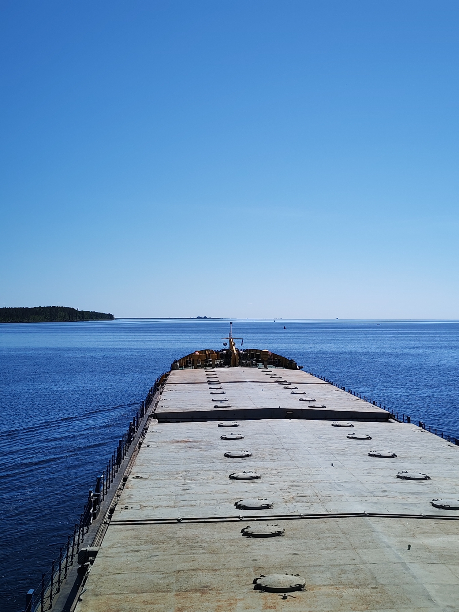 This image depicts the forward view from a large cargo vessel on a wide body of water, identified as being in the vicinity of Kostino, Russia. The scene is dominated by a clear, bright blue sky occupying the upper third of the frame. Below the sky is an expanse of deep blue water, showing ripples on the surface, particularly a distinct wake along the port side of the vessel, indicating forward motion.
The vessel's foredeck occupies the lower half to third of the image. It is a flat, expansive, light grey surface, appearing industrial and weathered. Numerous raised, oval or circular metal hatches, approximately 25-30 in number, are systematically arranged across the deck, suggesting access to cargo holds. A dark metal railing with vertical stanchions runs along the port side of the deck, extending towards the bow. At the very front of the visible deck, a small yellow-colored superstructure with industrial equipment, possibly winches or cranes, and a mast-like structure are discernible.
In the distance, on the left side of the horizon, a substantial landmass covered in dense, dark green trees is visible. Further to the right along the horizon, a fainter, low-lying land or structure is present. A faint, elongated horizontal structure, possibly a bridge or distant coastline, can be observed across the far horizon line. On the water in the middle distance, a red buoy is clearly visible, and several small, dark, indistinct shapes (likely other vessels or buoys) are scattered across the water towards the horizon. No people are visible on the deck or in the surrounding environment. The scene is under bright daylight conditions, indicated by direct sunlight and relatively short shadows on the deck hatches.