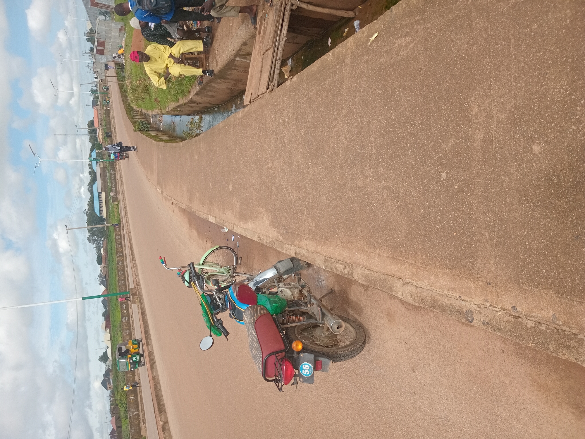 The image depicts a street scene in Bukuru, Nigeria, likely during daytime given the lighting. A paved road curves to the left, bordered by a concrete sidewalk on the right. A motorcycle and a bicycle are parked on the sidewalk near the road. Several people are visible, some walking on the road and others sitting or standing on the sidewalk. One man in a yellow outfit and red cap is seated on a wooden stool on the sidewalk. Another person in a red cap is walking on the road. In the background, there are buildings, trees, and a cloudy sky with multiple streetlights visible. A small, dirty canal runs alongside the sidewalk. The overall impression is that of a typical urban street in Nigeria, with evidence of informal settlements and local transportation.