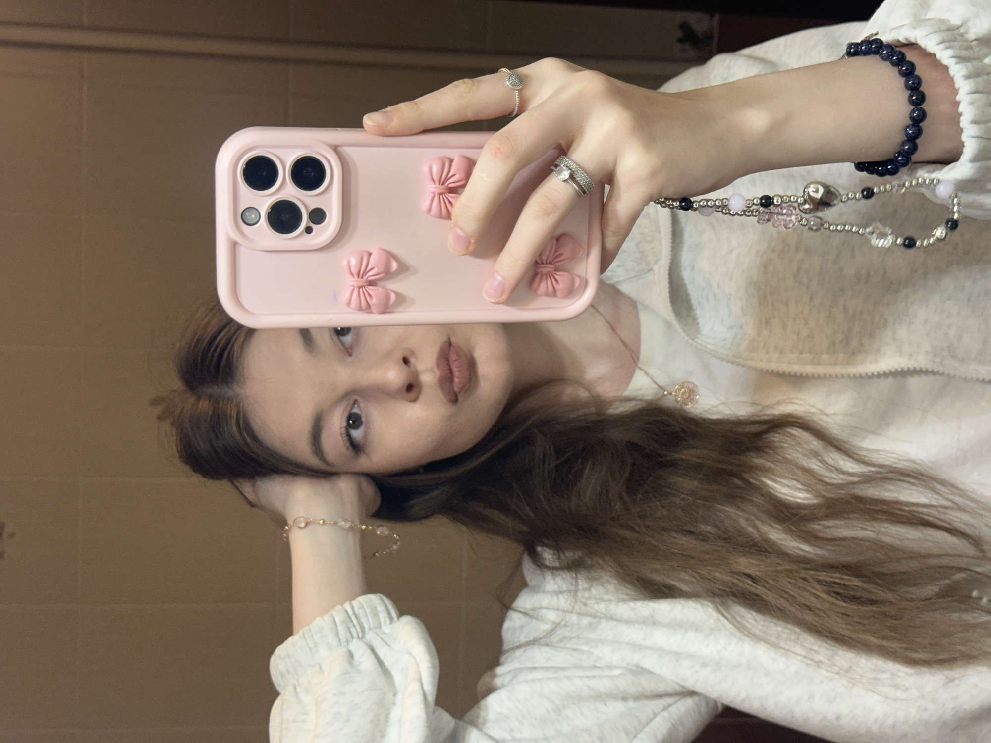A young woman is holding a pink phone case with three dimensional pink bows, while taking a selfie. The phone case features a triple-lens camera setup. She wears a ring on her pinky and ring finger. A bracelet with blue stones and a charm bracelet with beads, pearls, and a heart hang from the phone. She is wearing a white zip-up hoodie. Her hair is wavy and light brown. She is wearing a bracelet on her left wrist as well. The background is a blurred, light brown wall. The location appears to be indoors, likely a home, but is not specified within Kazan, Russia.