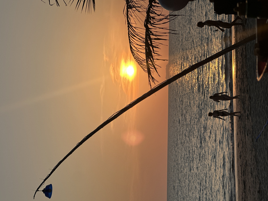 This image captures a sunset over a beach in Phra Ae, Thailand. The horizon shows a setting sun, creating a bright orange and yellow sky with a reflective path on the ocean surface. The ocean exhibits calm conditions with small waves reaching the sandy beach. A tall, dark pole with a blue, downward-facing light fixture at its apex extends diagonally from the lower left towards the upper right. Silhouetted palm fronds are visible on the right side of the frame. Three human figures are silhouetted on the sandy beach near the waterline. Two figures, presumed female and wearing swimwear, walk from left to right. A third figure, presumed male and wearing swimwear, walks from right to left, positioned further right. A faint, dark outline consistent with a distant island or large vessel is present on the horizon, directly in line with the sun's reflection. A partially visible, blurry red and yellow object, possibly a container, occupies the extreme lower right foreground.