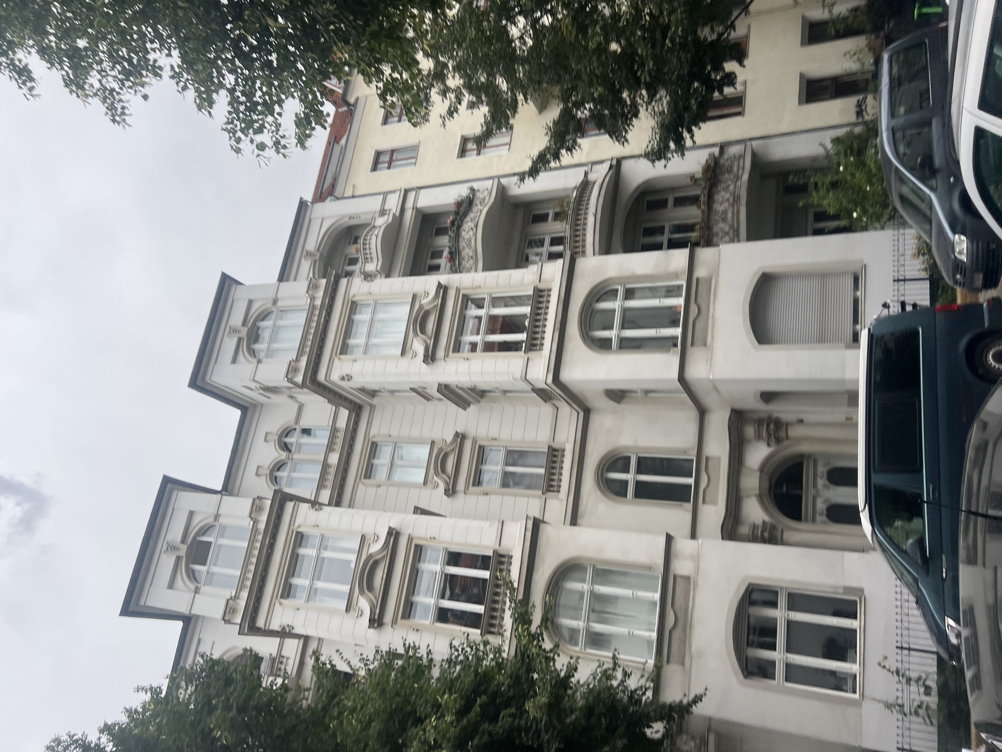 The image shows a street view in Berlin, Germany. A white apartment building with a pale yellow adjacent building dominates the scene. The white building features multiple stories with numerous windows, some arched and some rectangular, adorned with decorative moldings. Several balconies, some with ornate railings and flower boxes, are visible on the facade. The yellow building has red-tiled roofs and visible window frames. Trees with green foliage partially obscure the buildings on the left and right sides of the frame. In the foreground, several vehicles are parked or driving on the street. A dark green van is prominently positioned in the lower left, and parts of a dark gray and a white car are also visible. A black metal fence runs along the sidewalk in front of the buildings. The sky is overcast and appears cloudy.