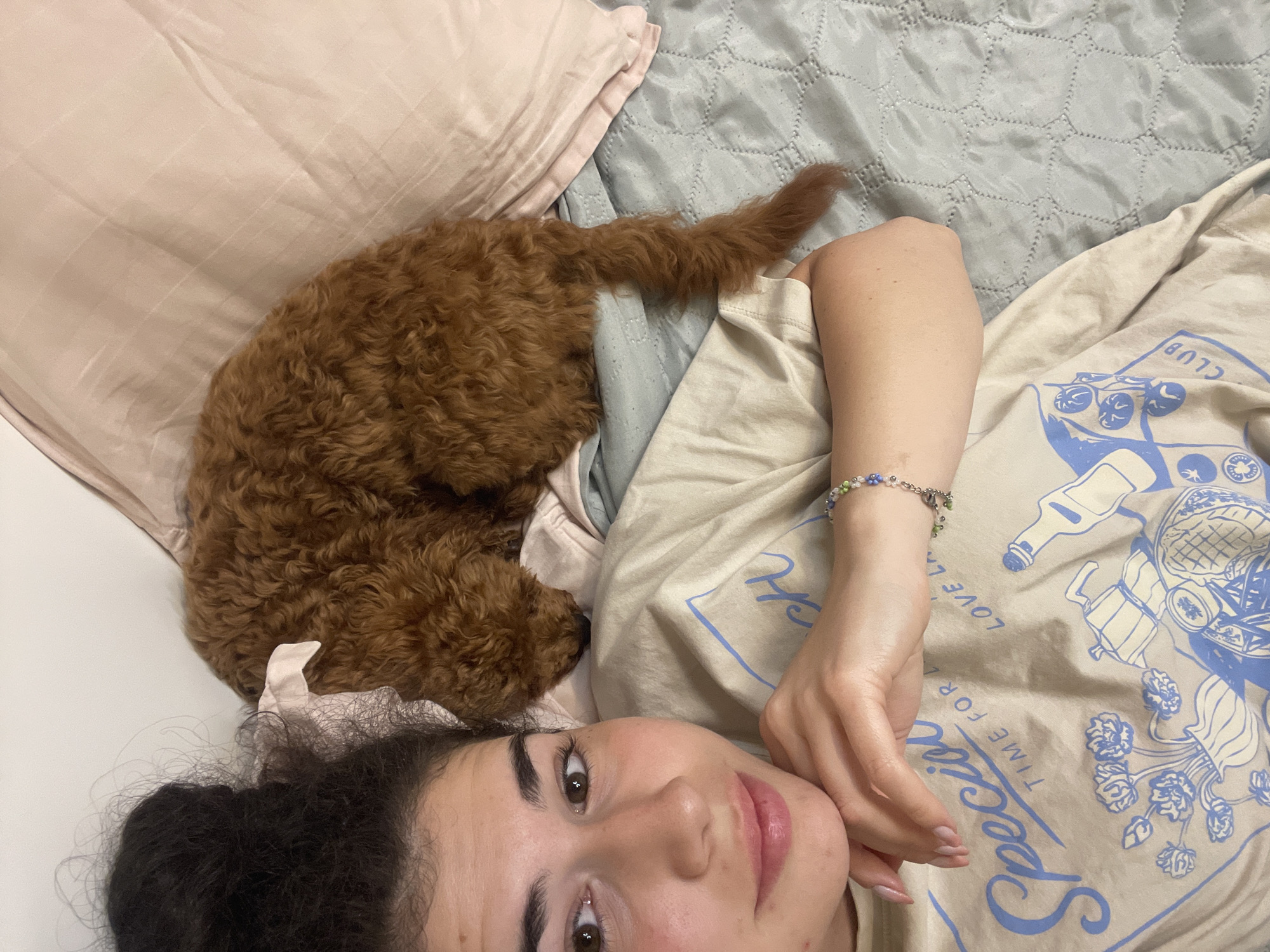 A young woman with dark hair pulled up in a messy bun is lying on her side, looking at the camera. She is wearing a beige t-shirt with a blue graphic print. Her left arm is resting on her chest, and her hand is supporting her chin. She is wearing a bracelet with colorful beads on her left wrist. A small, reddish-brown curly-haired dog is sleeping next to her, with its head tucked in. The dog is wearing a small pink bow on its head. They are lying on a bed with a light blue textured comforter and a pale pink pillow. The setting appears to be a bedroom. There are no visible indicators that this scene is located in the city of Sofia, Bulgaria.