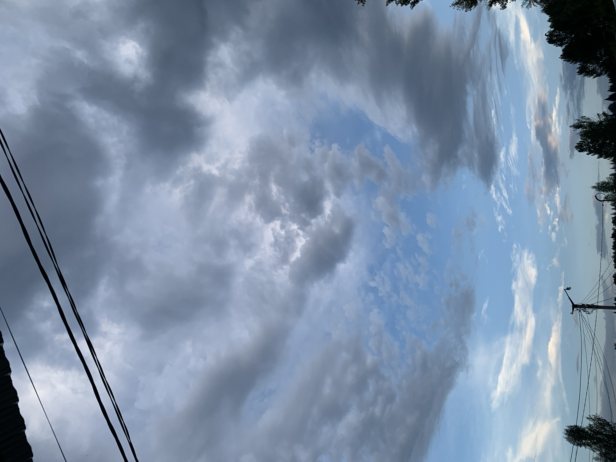 The image captures a dynamic sky over Asha, Russia, featuring a dramatic interplay of dark, heavy clouds and patches of bright blue sky. Two black electrical wires are prominently strung across the upper portion of the frame, with a hint of a corrugated metal roof visible in the top left corner. Below the wires, a landscape unfolds with silhouetted trees and a utility pole with a light fixture. The scene suggests a potentially stormy or overcast day, with the rugged texture of the clouds hinting at changing weather conditions.