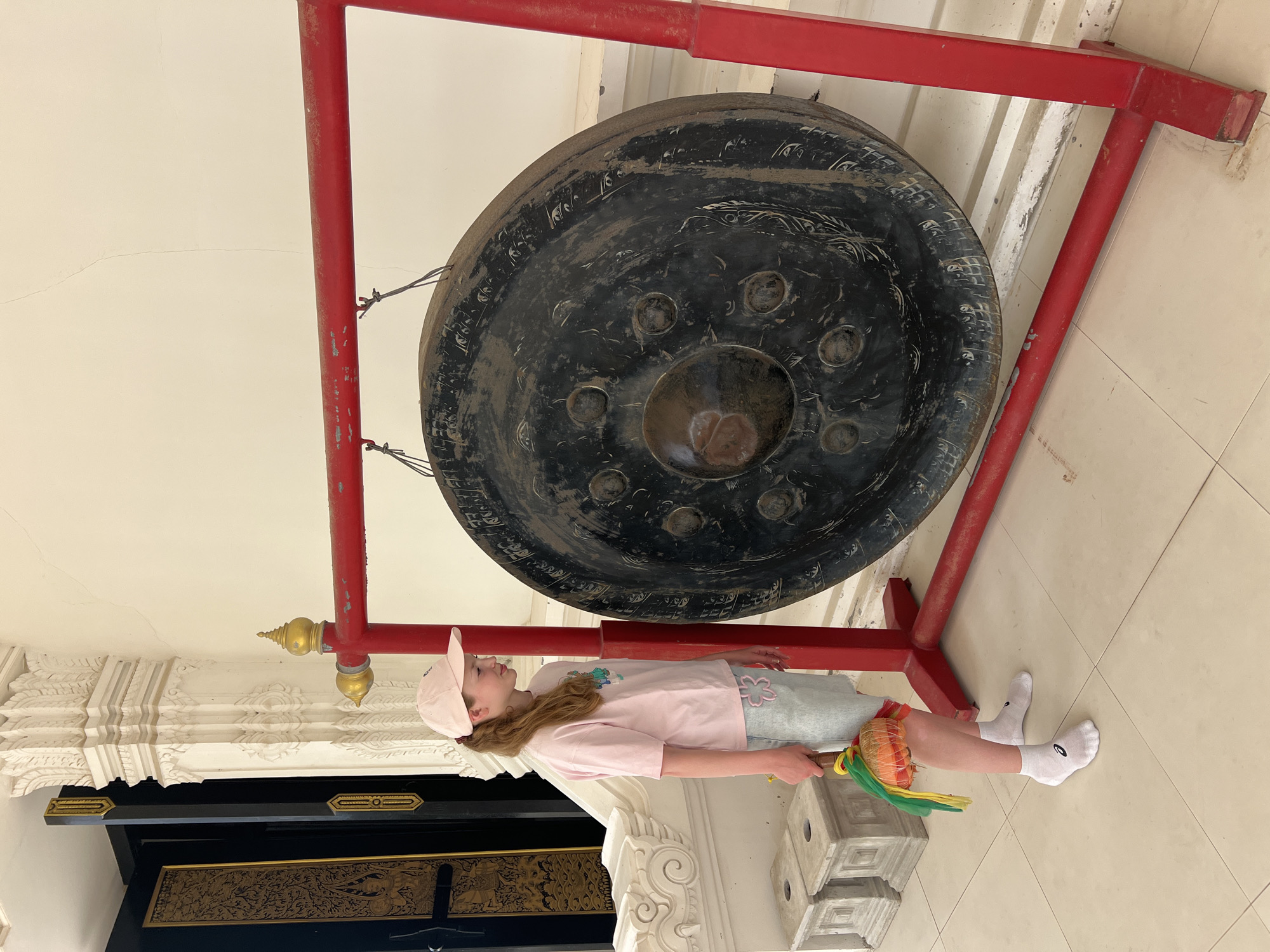A young person wearing a pink baseball cap, a pink t-shirt, and light blue shorts stands to the left of a large, dark gong. The gong is suspended from a red metal frame. The person holds a mallet with a colorful cloth in their right hand. The background features ornate white architectural elements and a dark, decorated doorway. The scene appears to be outdoors, with light-colored tile flooring. The location is identified as Baan Bo Phut, Thailand.