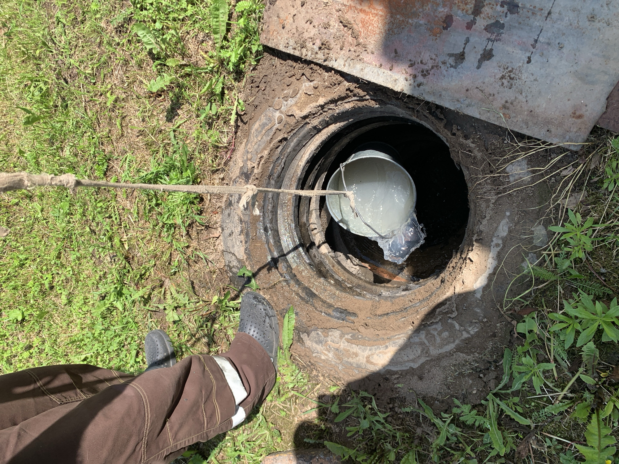 A person wearing brown pants with reflective strips on the lower legs and dark gray clogs stands near an open manhole. A bucket filled with a light-colored liquid is suspended by a rope, partially lowered into the manhole. The surrounding ground is covered in grass and dirt, with a rusty metal plate visible to the right of the manhole. The scene appears to be outdoors, likely in a rural or suburban area.